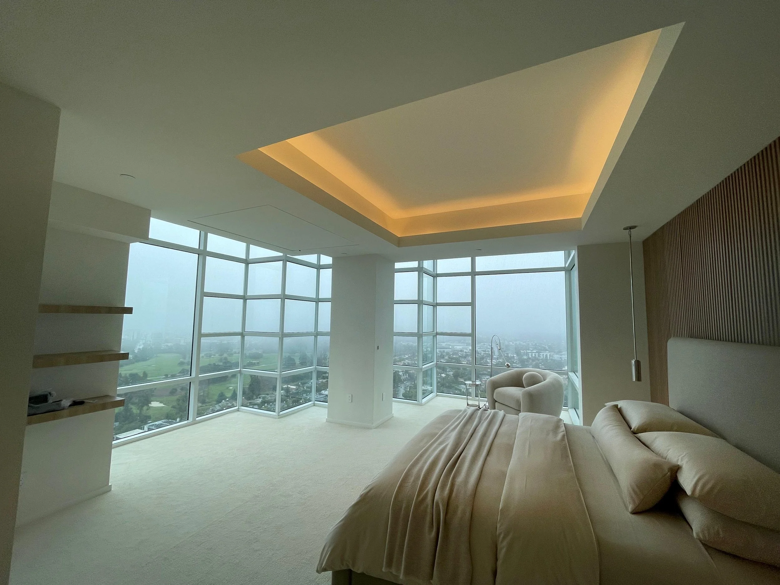 Modern living room with large floor-to-ceiling windows, a beige sofa, a hanging pendant light, and a view of a cityscape and green landscape outside.