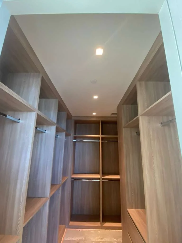 A walk-in closet with wooden shelves and hanging rods, illuminated by ceiling lights.