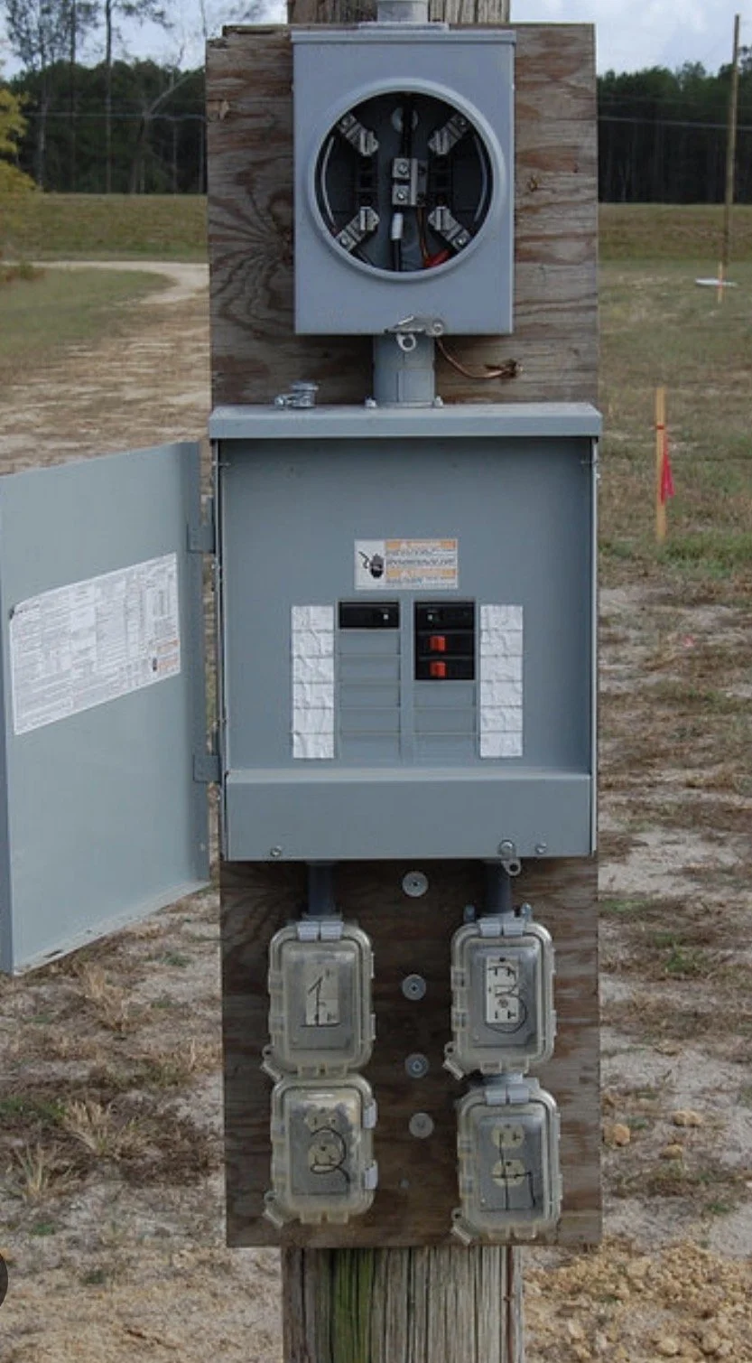 An electrical meter box on a wooden post outdoors with meters and circuit breakers visible, set against a grassy area and power lines.