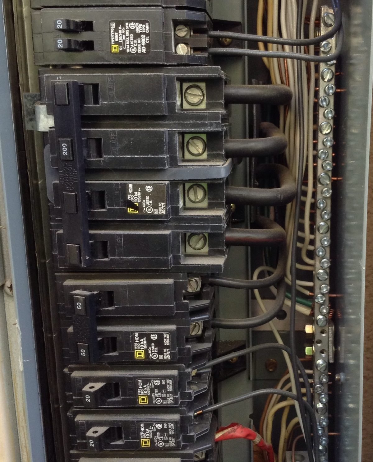 Close-up of electrical circuit breaker panel with multiple black breakers, connected to wires and metal bus bar.