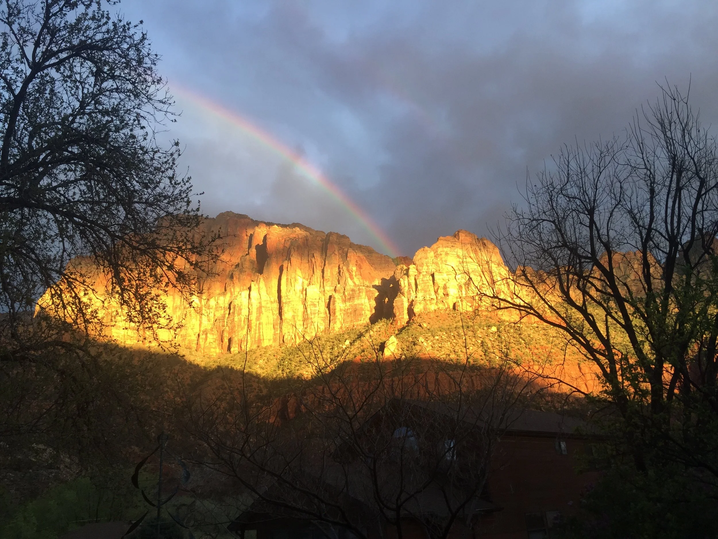 Sunlight illuminating a mountain range with a rainbow in the sky above, trees in the foreground, and a cloudy sky.