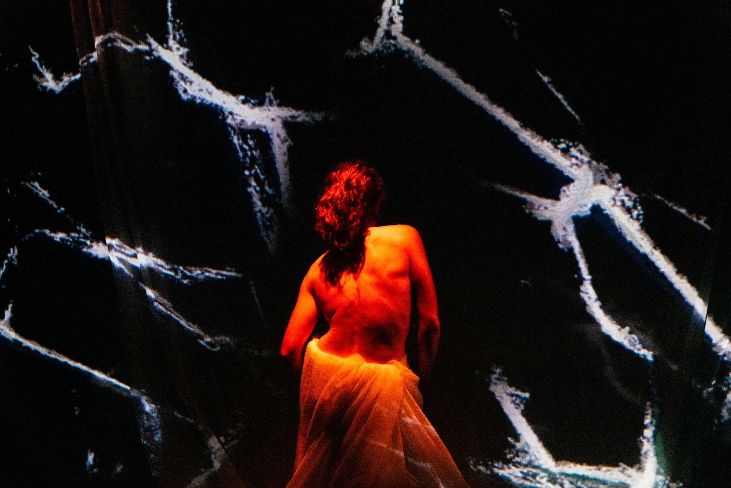 A person with long, curly hair and a bare back, wearing a flowing skirt, stands in front of a large digital screen displaying abstract white and blue lines on a black background.