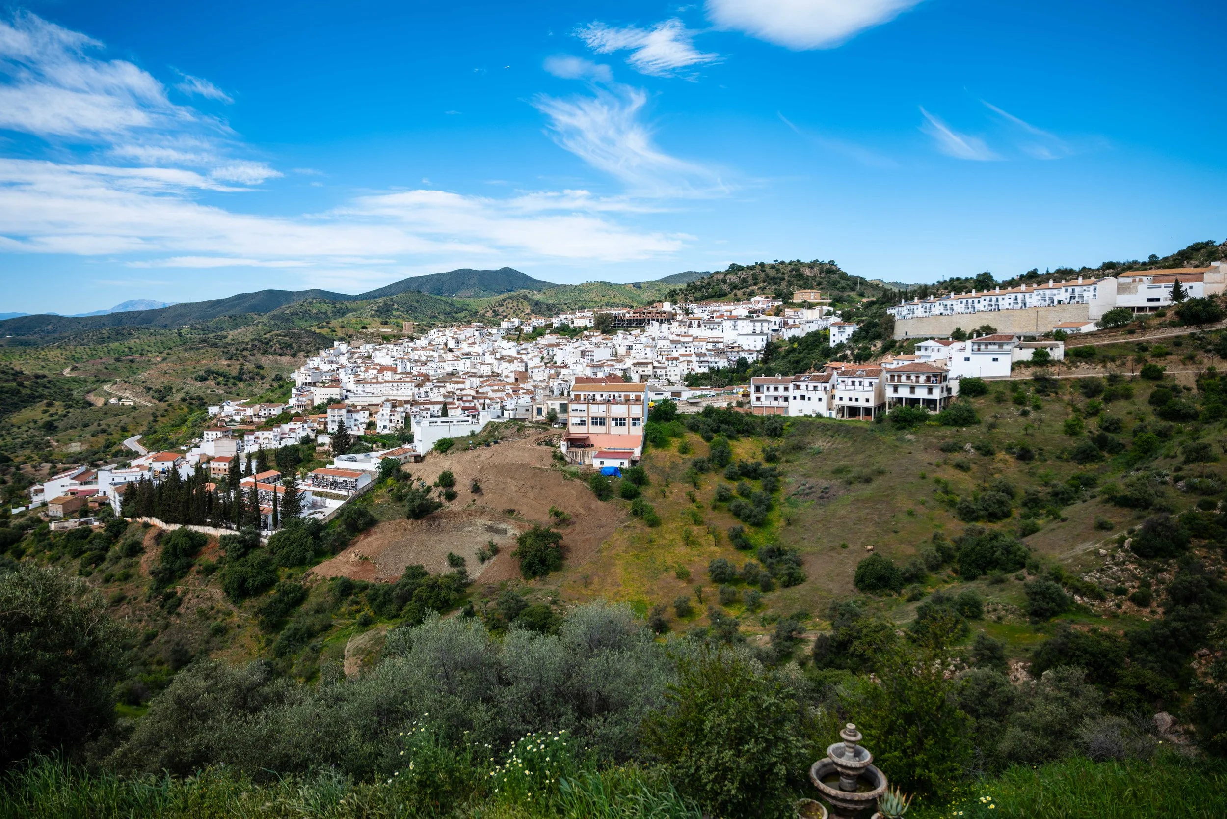 A hillside town with white buildings and terracotta roofs nestled among green vegetation under a blue sky with scattered clouds.
