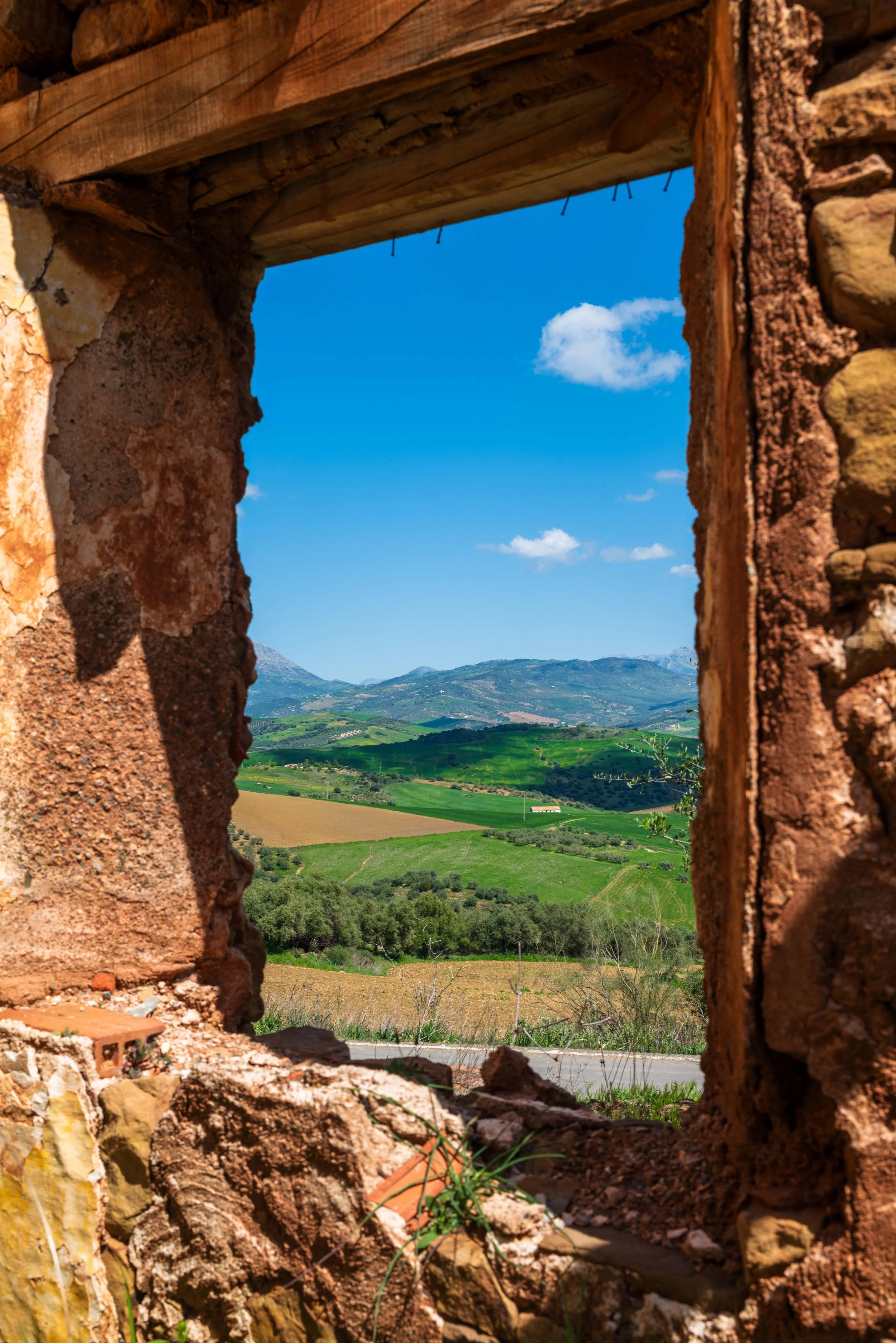 View of rolling green hills and mountains through an old, crumbling stone window frame under a bright blue sky with a few white clouds.
