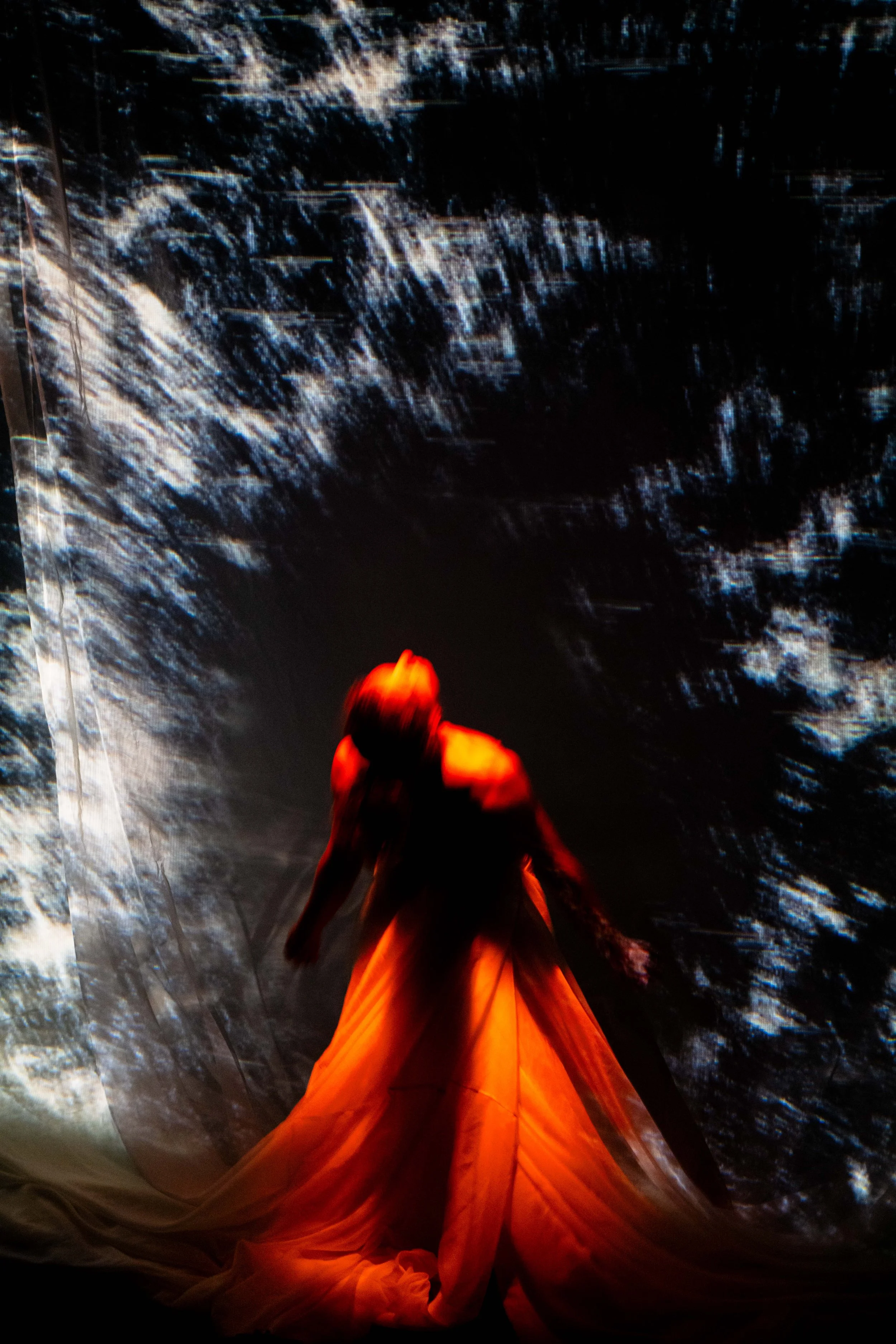 A person in a flowing orange dress standing under a large abstract projection with dark blue and white streaks.