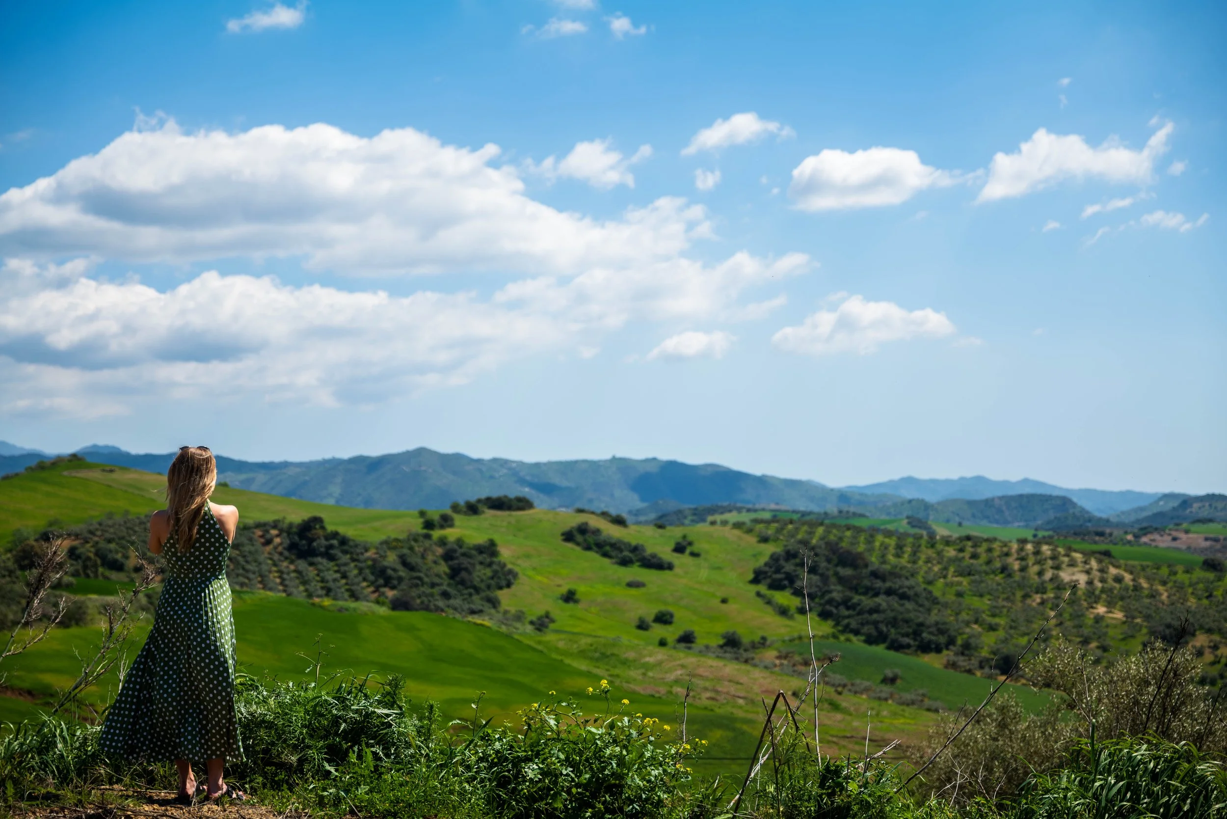 Woman in a polka dot dress standing on a hill overlooking rolling green hills and mountains under a blue sky with scattered clouds.