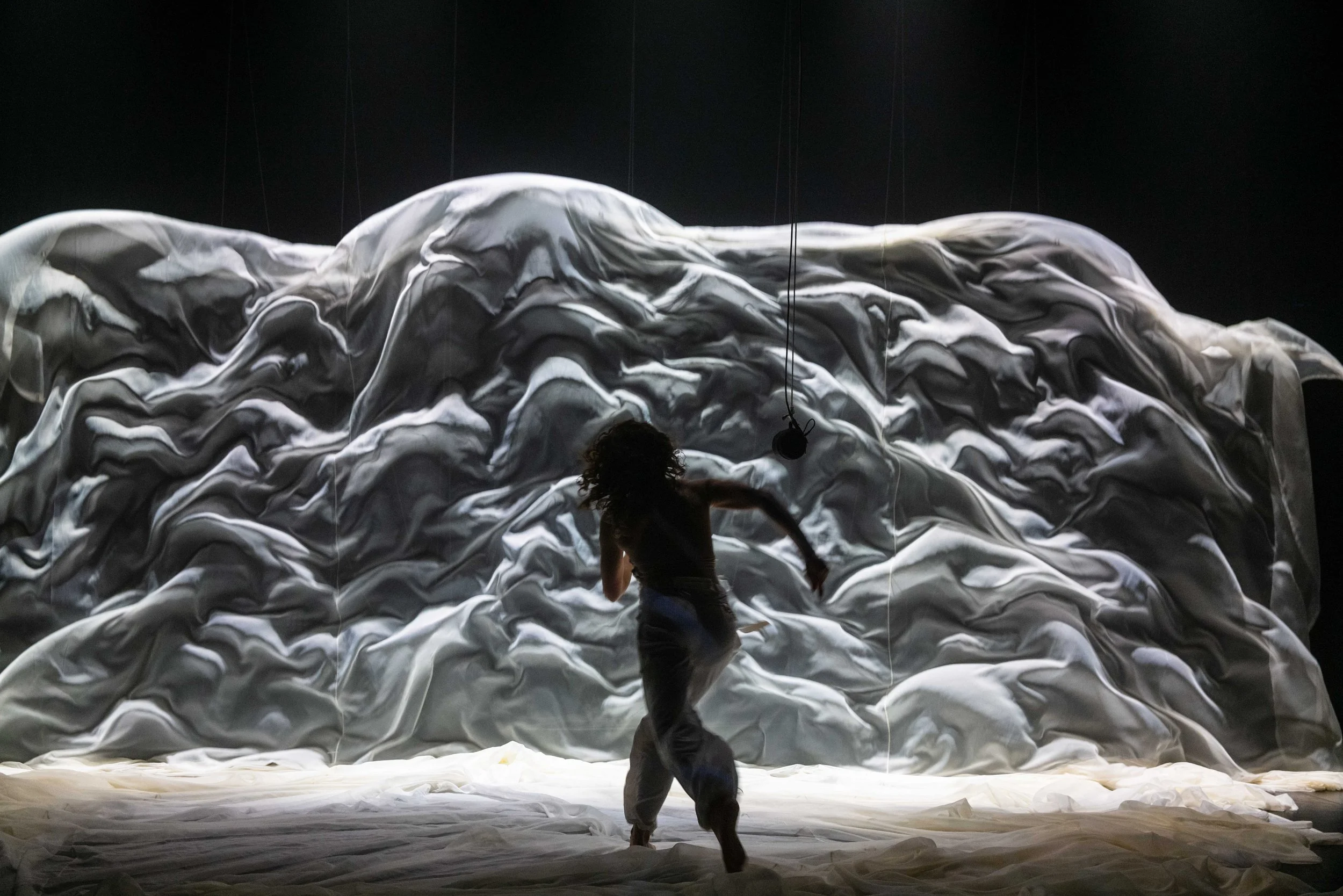 A person with long curly hair performing a dance or theatrical act in front of a large, abstract textured backdrop resembling a mountain or cloud formation, dimly lit against a dark background.