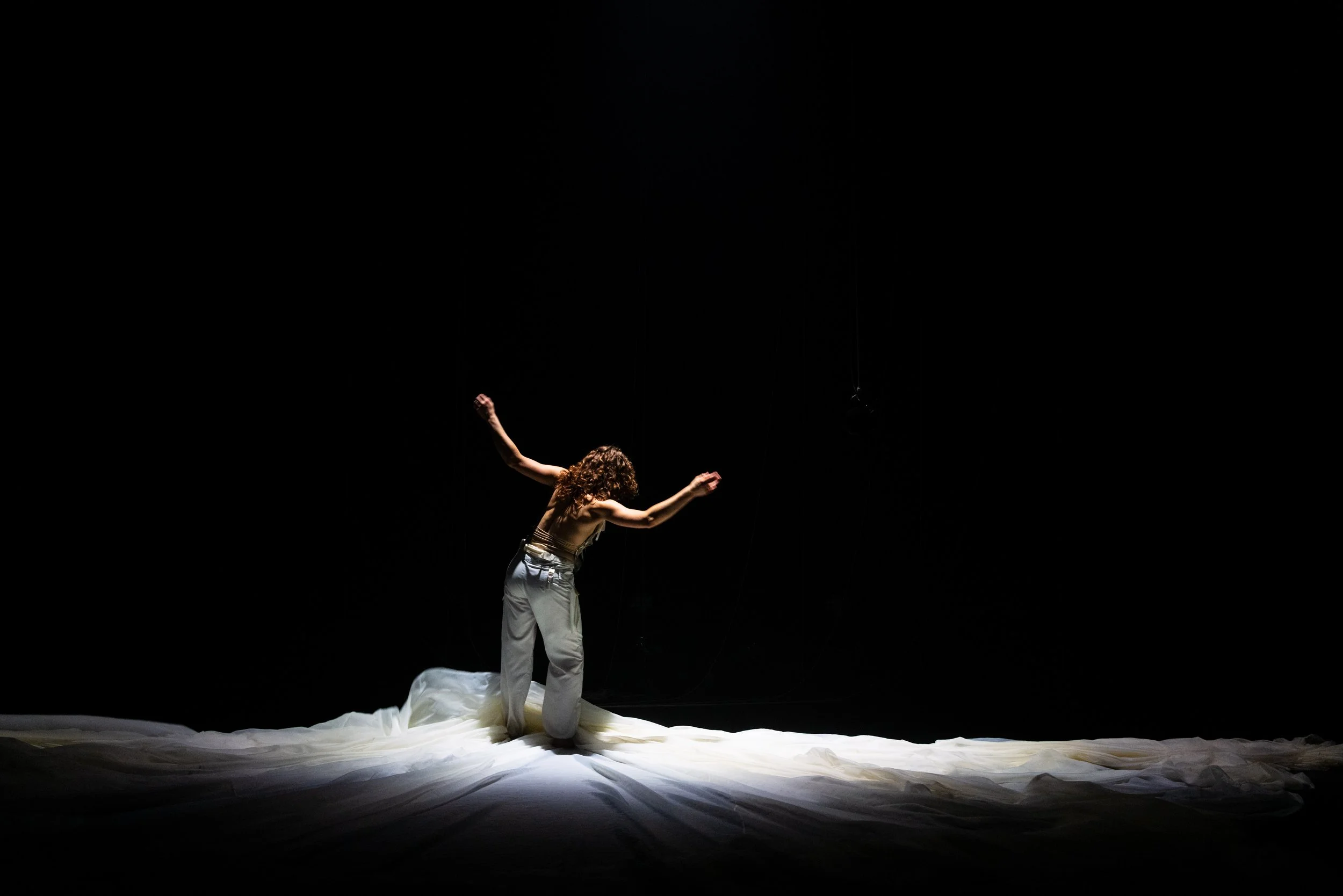 A woman with curly hair standing on a stage, illuminated by a spotlight, with a dark background and white fabric on the floor.