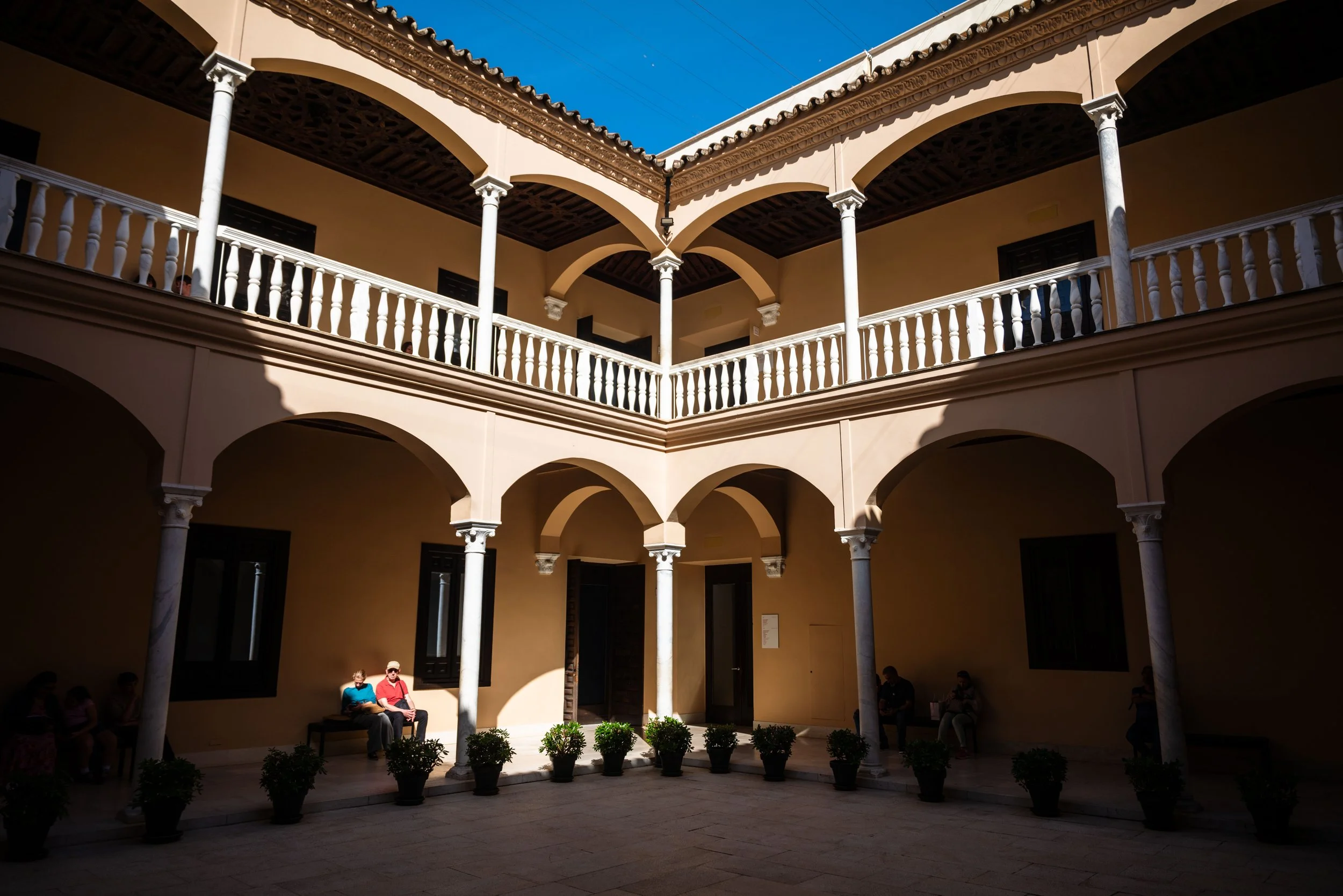 An interior courtyard of a historic building with two floors, arched walkways, white columns, and a balcony. There are potted plants along the ground and several people seated on benches and chairs in the shaded areas.