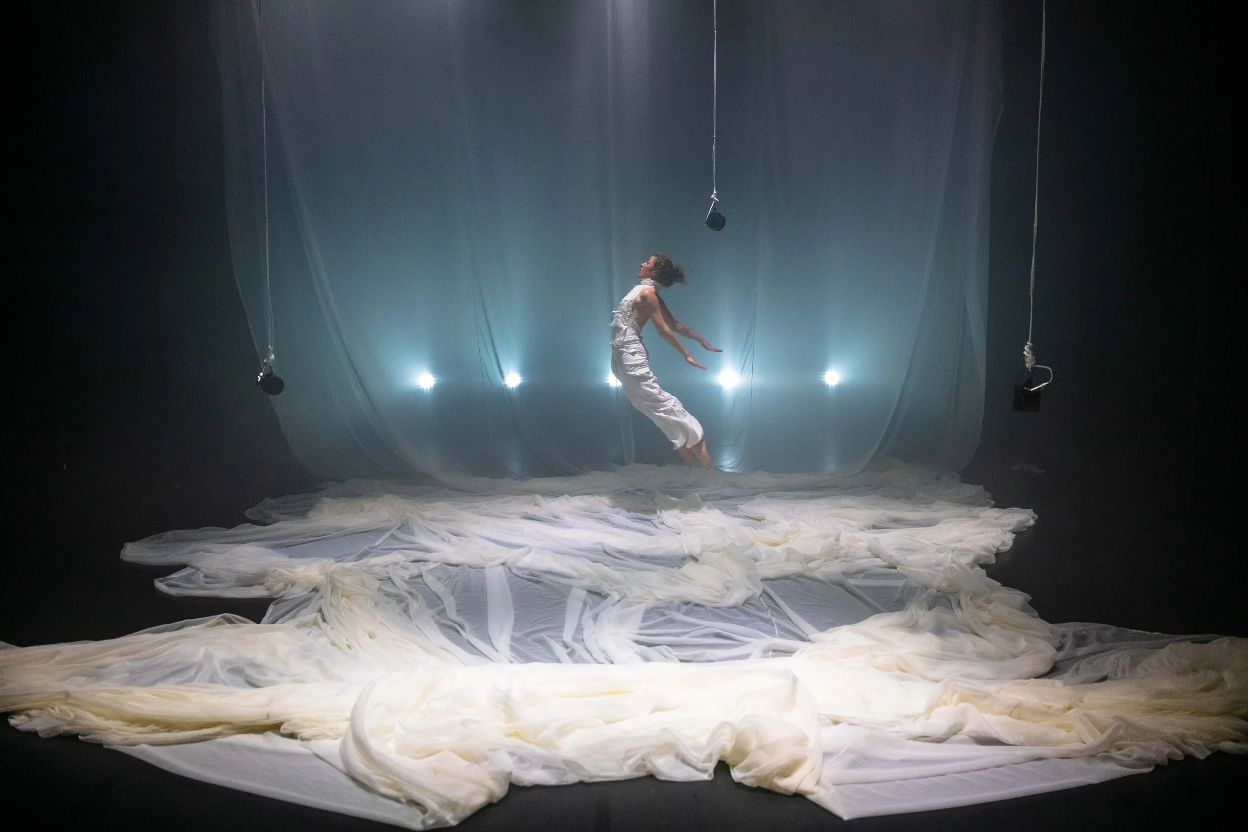 A woman in a white dress is jumping in the air on a stage with sheer fabric and fabric on the ground, illuminated by backlights, with hanging microphones around her.