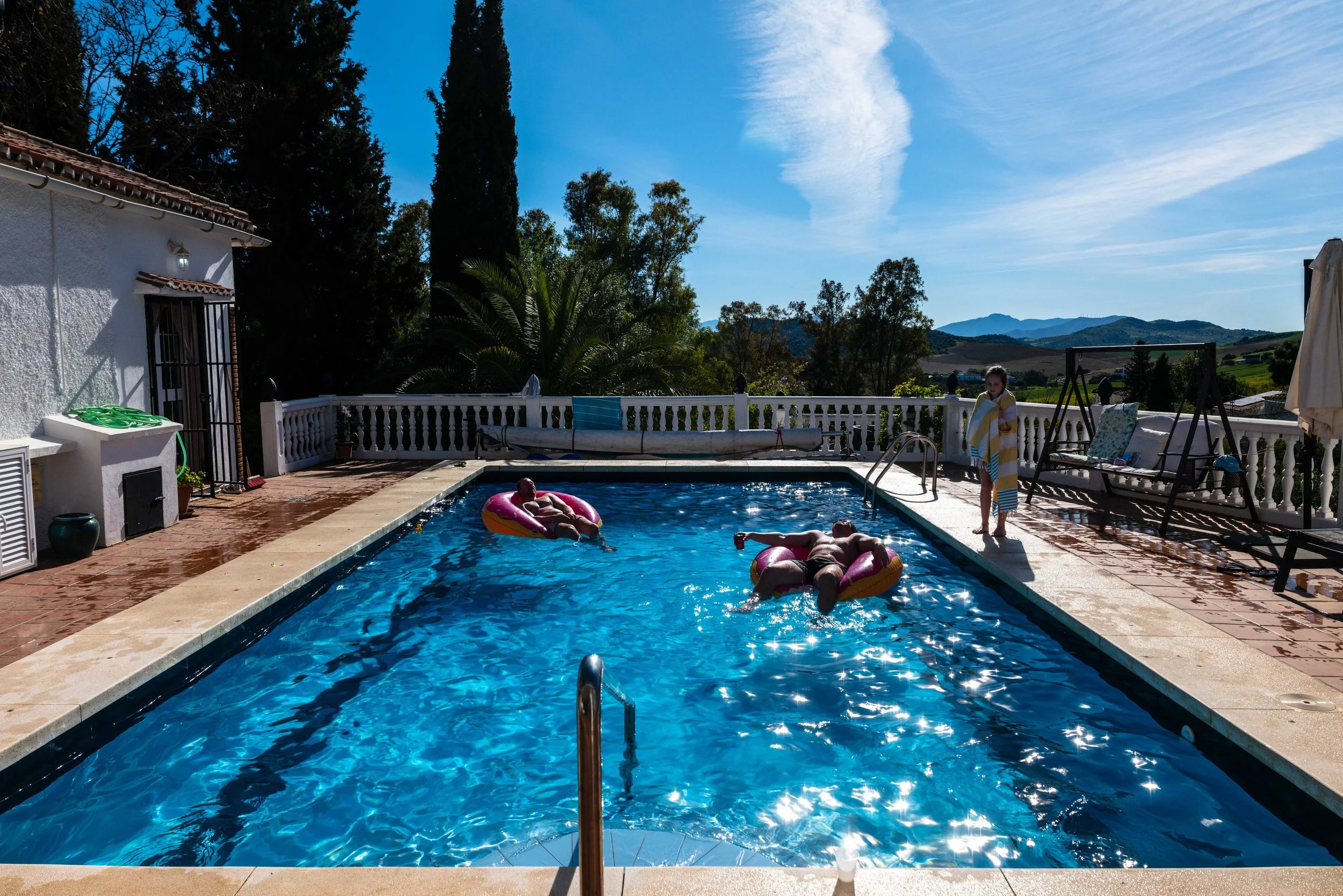 People relaxing in a backyard swimming pool on a sunny day, with mountains and trees in the background.