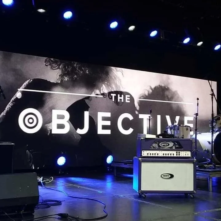 A blue hued image showing an empty stage at the Objective summit, Nashville. An amplifier, drum kit and cables represent the idea of supporting us with equipment, tech or other corporate sponsorship.