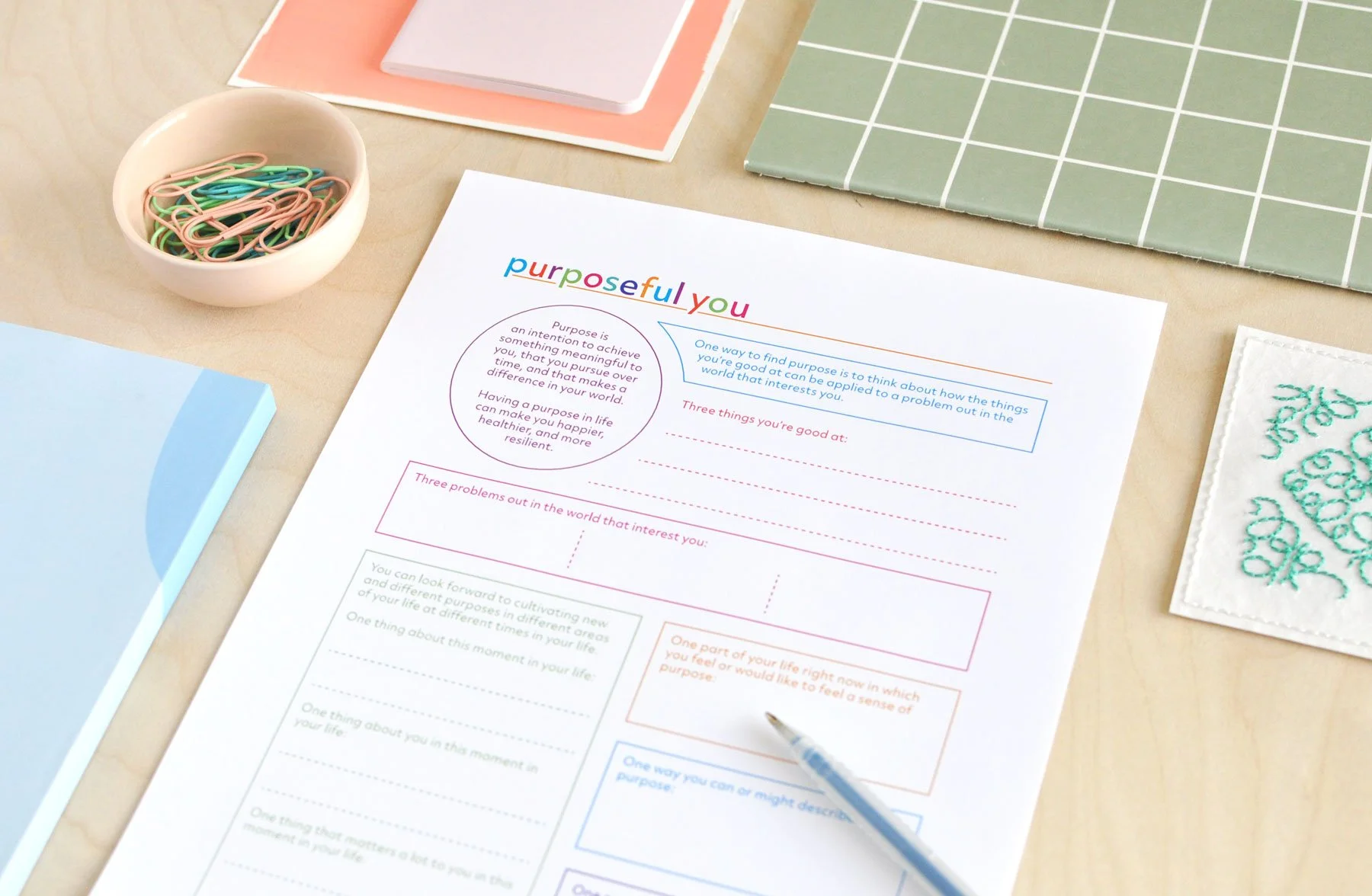 Colourful journal page titled Purposeful You filled with writing prompts for reflecting on your purpose. Shown with colourful notebooks and desk items on a light wood background.