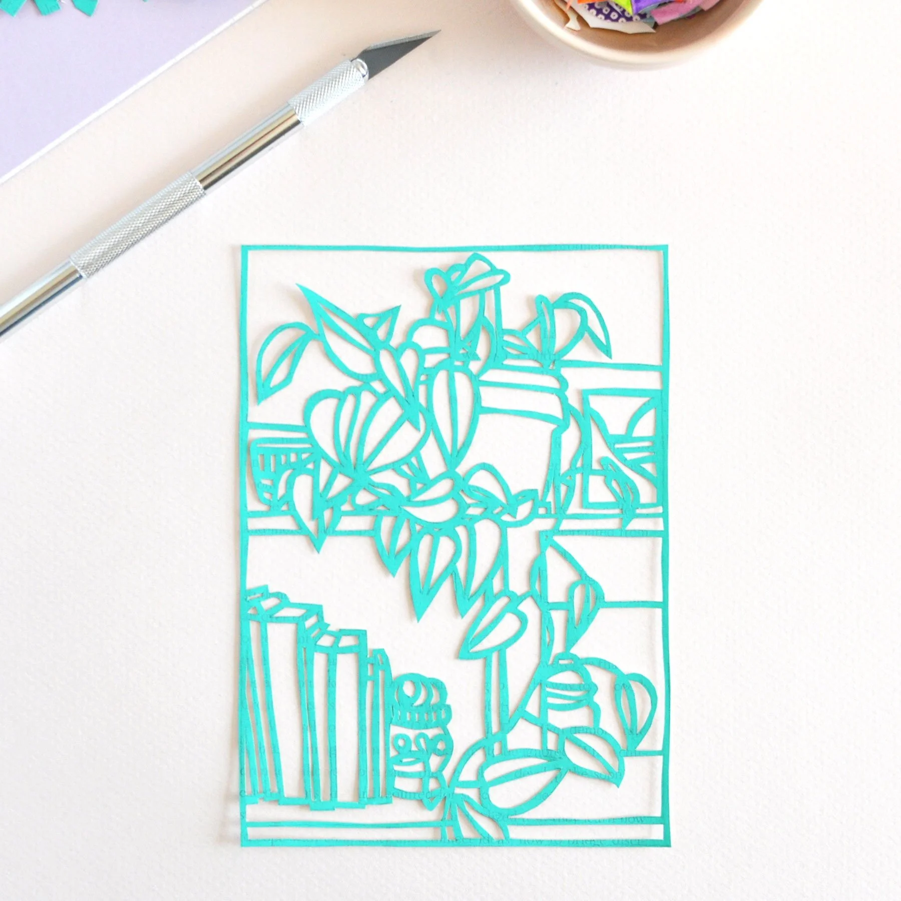 A small paper cutting of a plant on a bookshelf made from a book page painted green. Shown with an excacto knife and a glimpse of paper scraps.