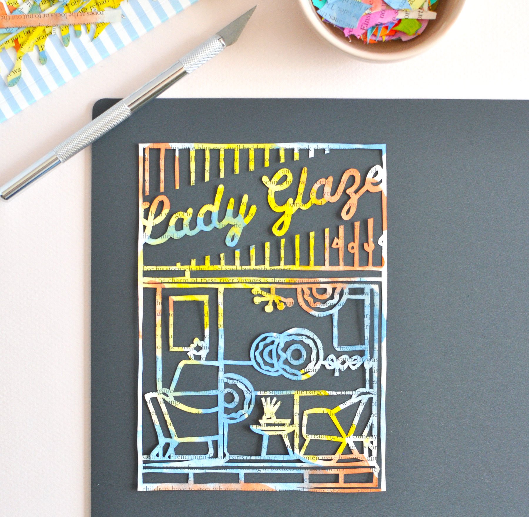 A colourful paper cutting of a donut shop storefront. Shown with craft knife and a small dish filled with colourful paper scraps.