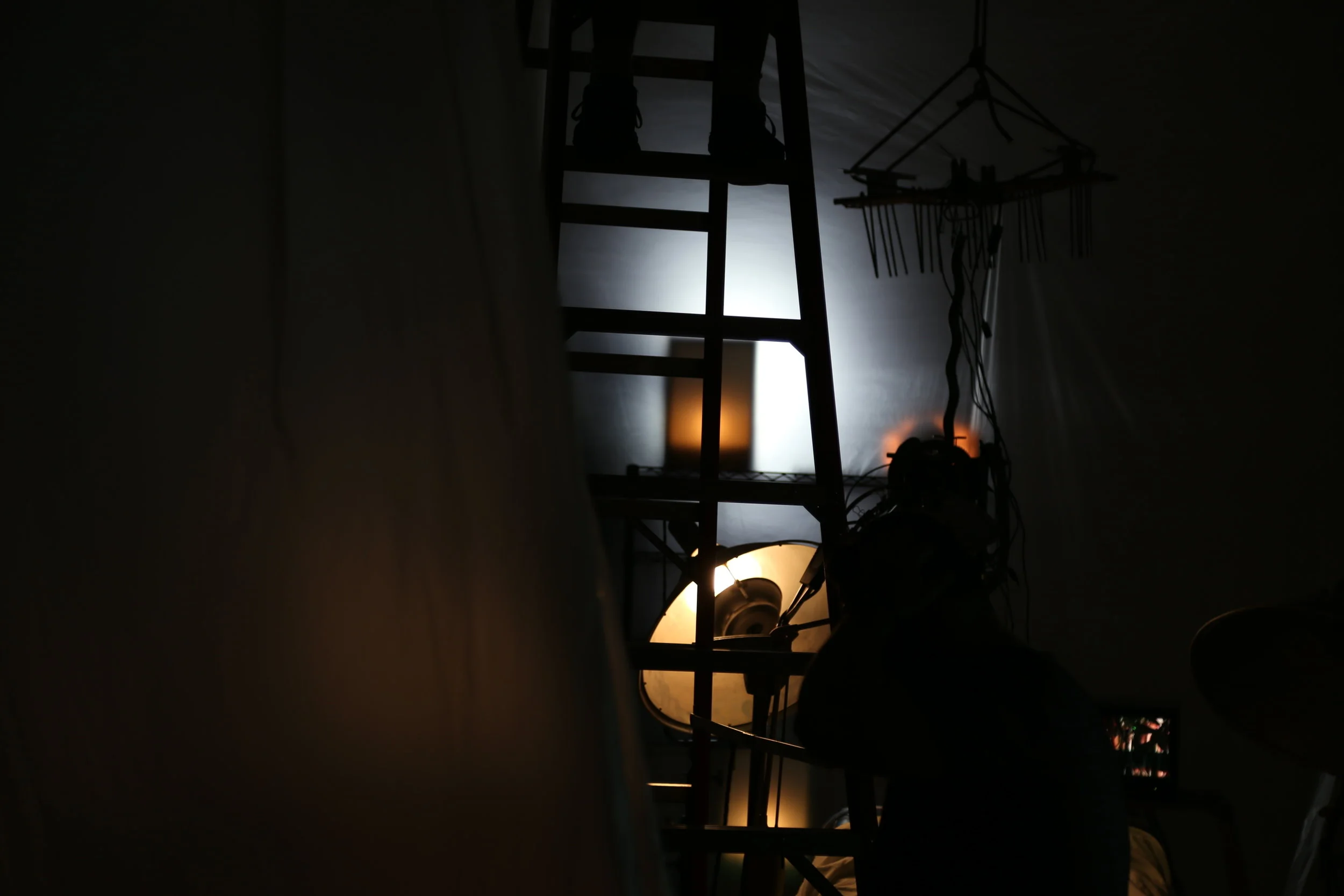 Dark indoor scene with a tall ladder, hanging lights, and various cables. The background includes a white wall and some illuminated equipment.