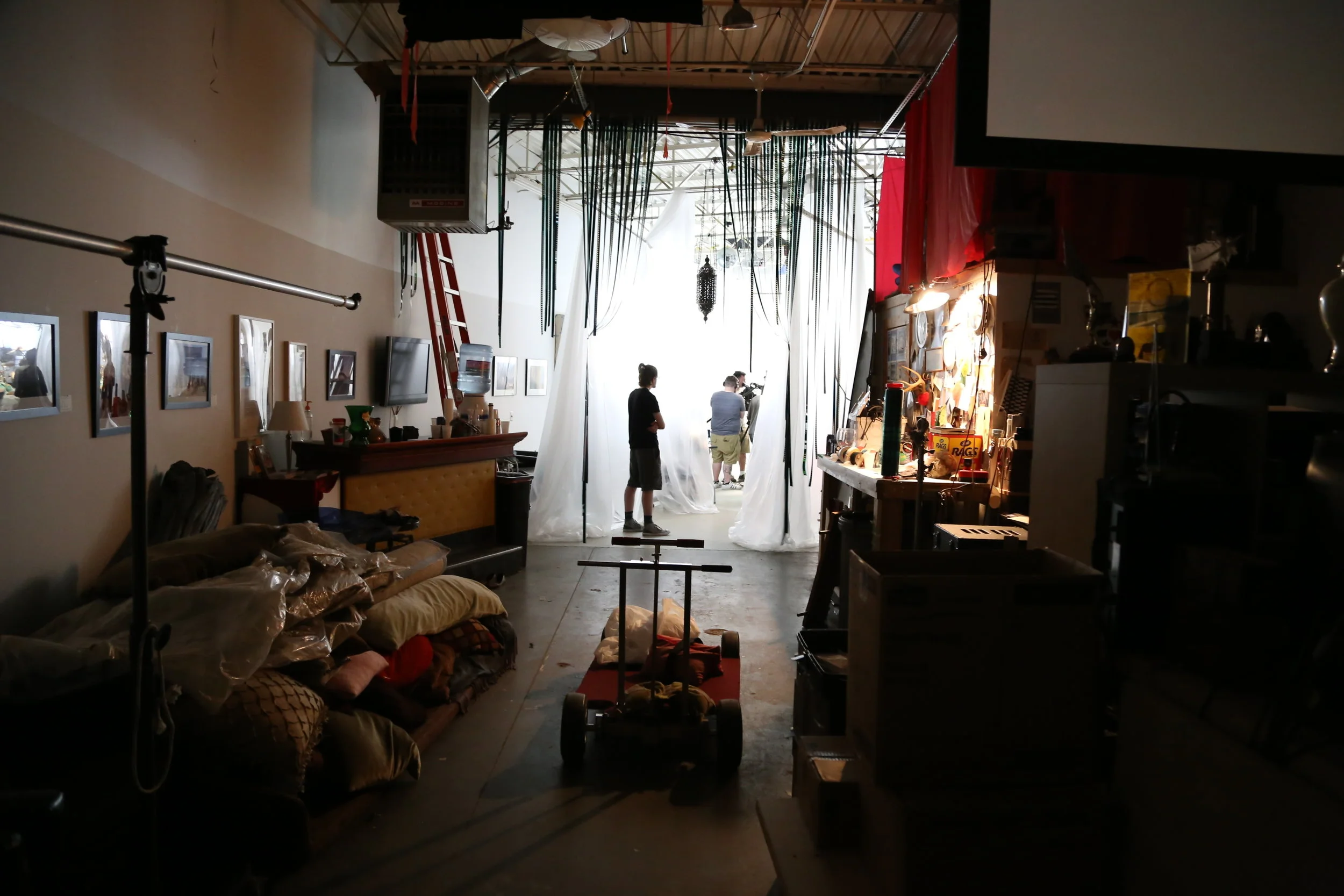 Inside a studio or workshop with various equipment and supplies, including a backdrop, cameras, lighting, and people working, with natural light coming from the back.