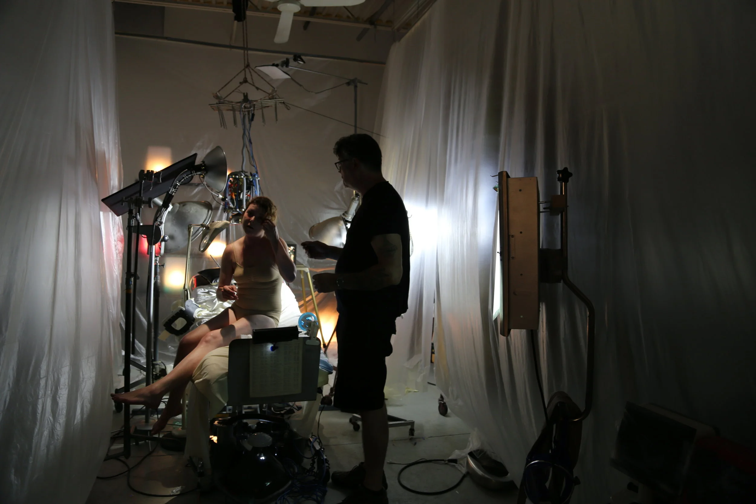 A medical or scientific setup inside a clinical or laboratory environment with a woman sitting on a medical bed and two men engaging in a discussion. The room is enclosed by white curtains and has specialized medical or technical equipment around.
