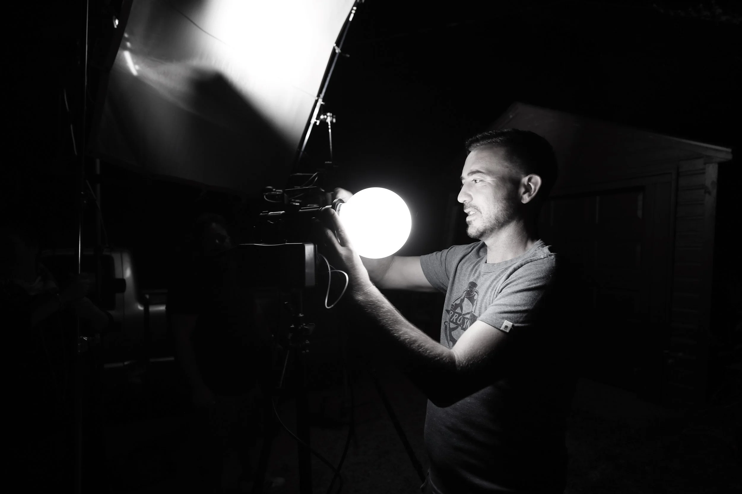 A man in a dark room interacts with a glowing spherical object, illuminated by a light, with studio equipment visible.