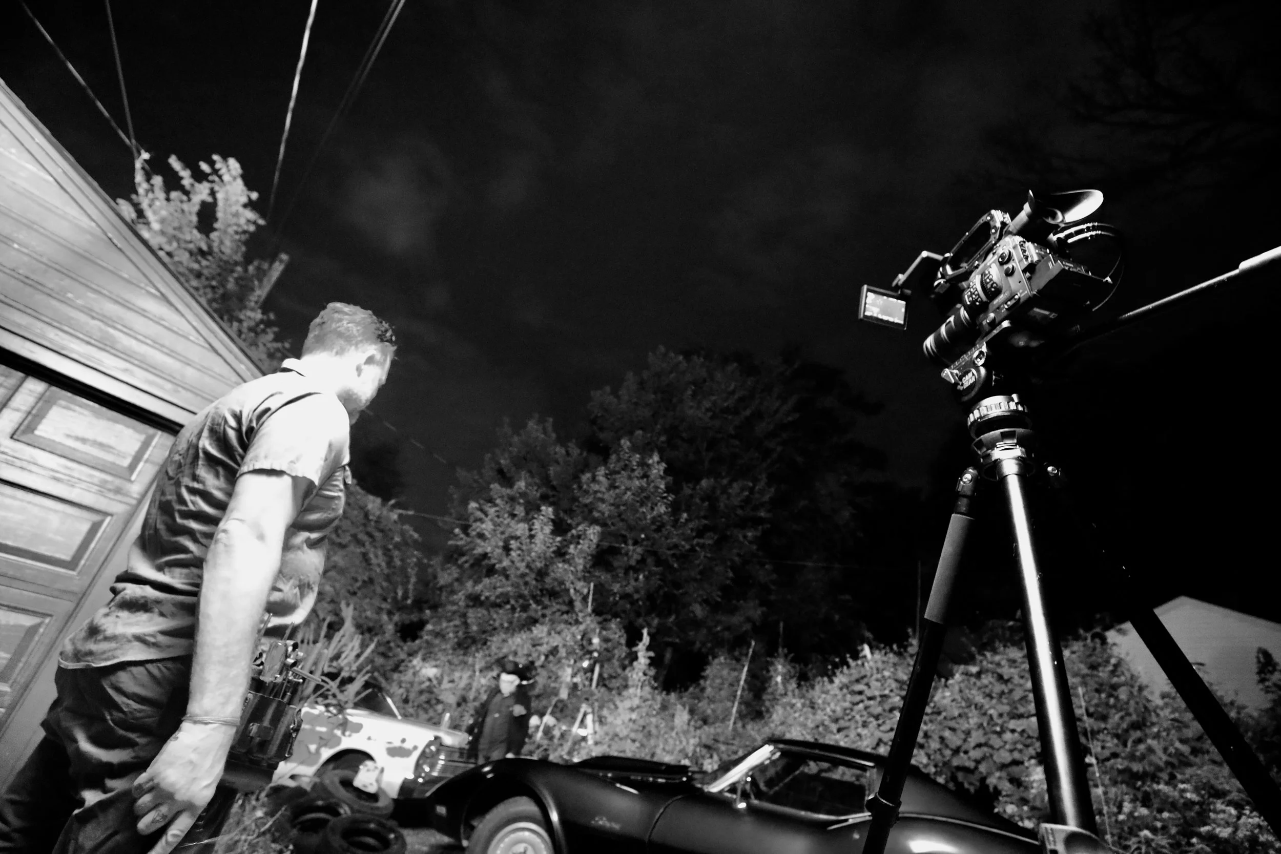 A person standing outside at night with a professional video camera mounted on a tripod, capturing the scene. The person is near a wooden structure, with trees and parked cars in the background under a dark sky.
