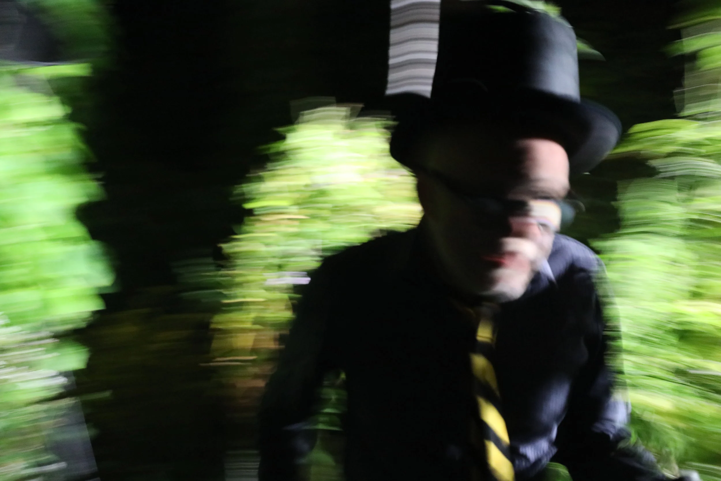 Person wearing a black hat and dark clothing, possibly wearing glasses, holding a camera or phone, surrounded by green foliage, with motion blur effect.
