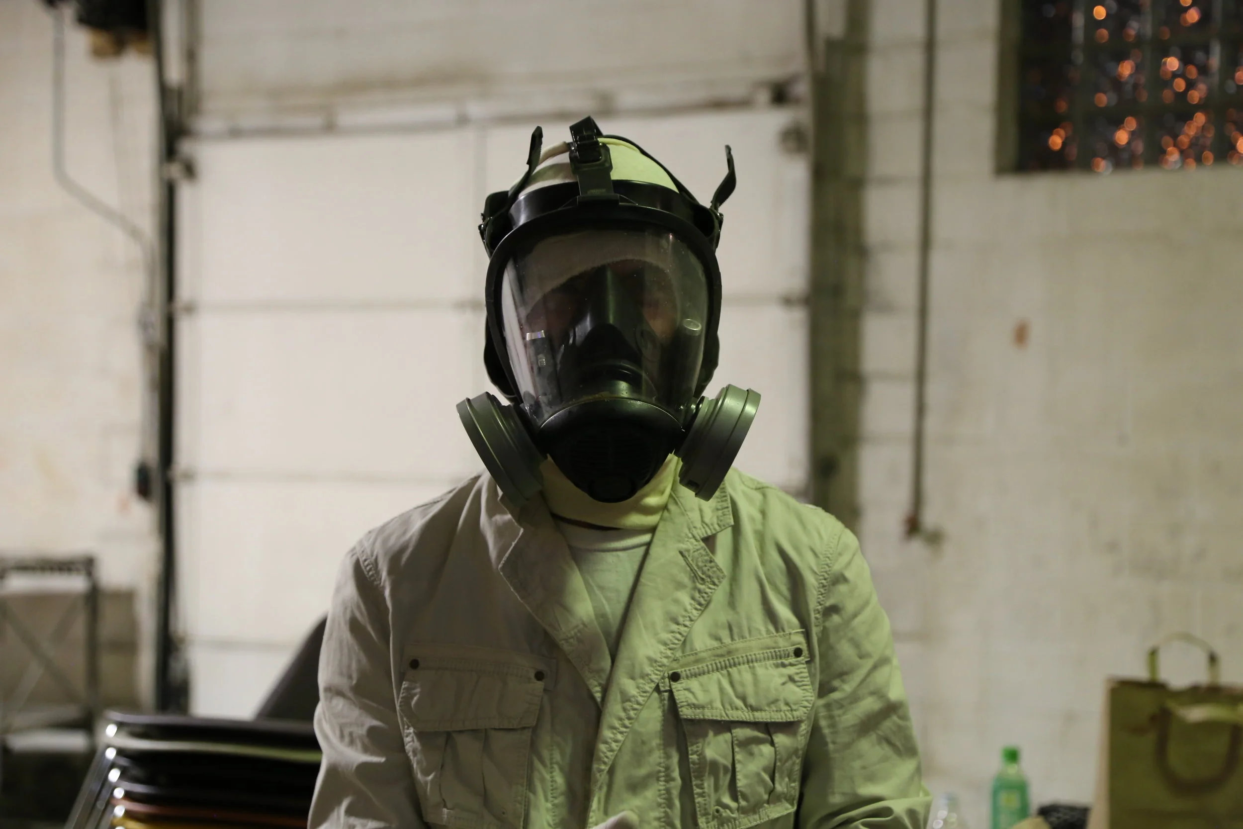 A person wearing a protective face mask with a visor and a beige jacket in an industrial or warehouse setting.