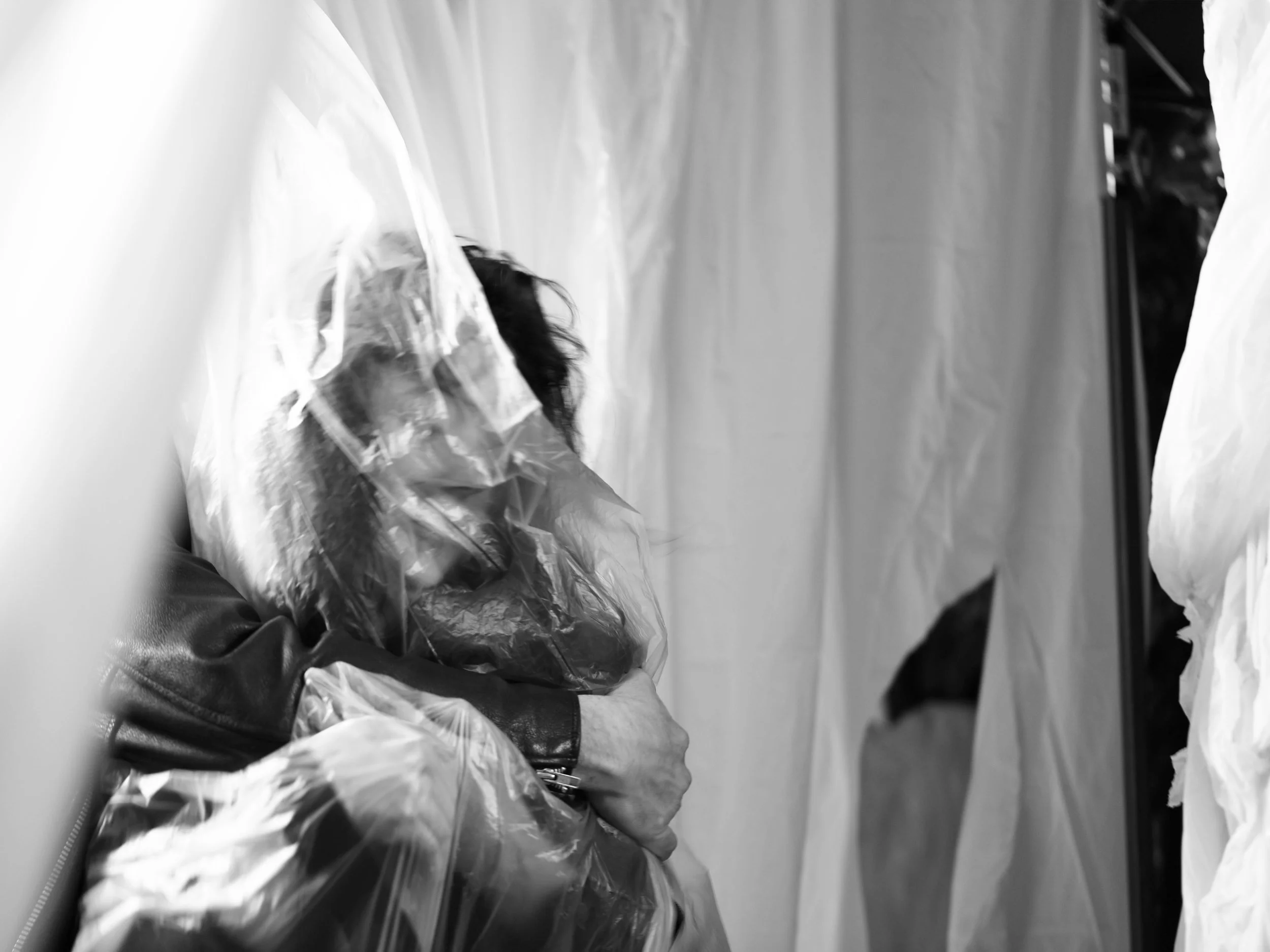 Black and white photo of a person sitting at a table with plastic bags around them, possibly in a setting like a store or warehouse.