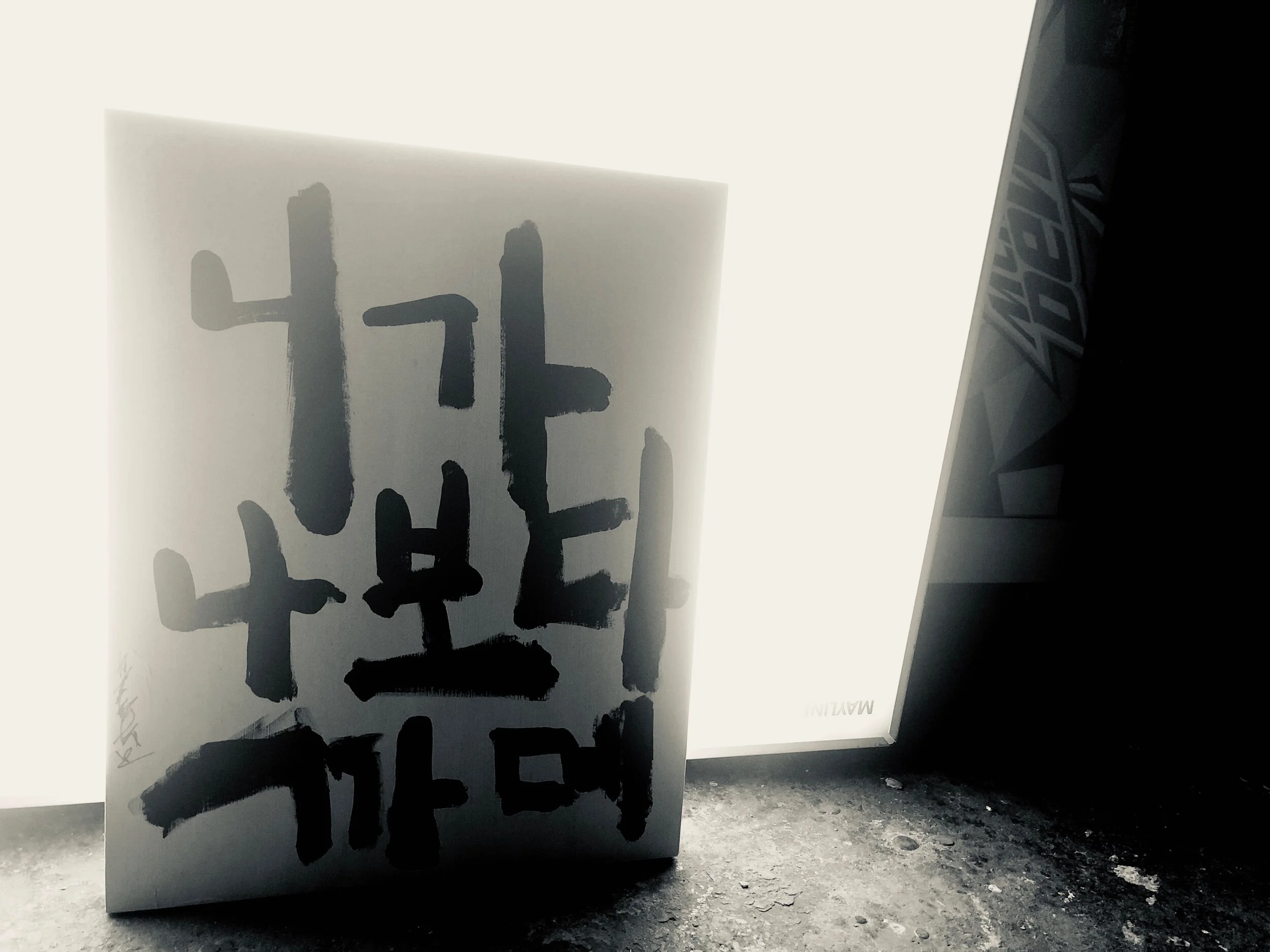 Close-up of a white poster with black abstract Japanese calligraphy characters, leaning against a wall with a small gap revealing the ground.
