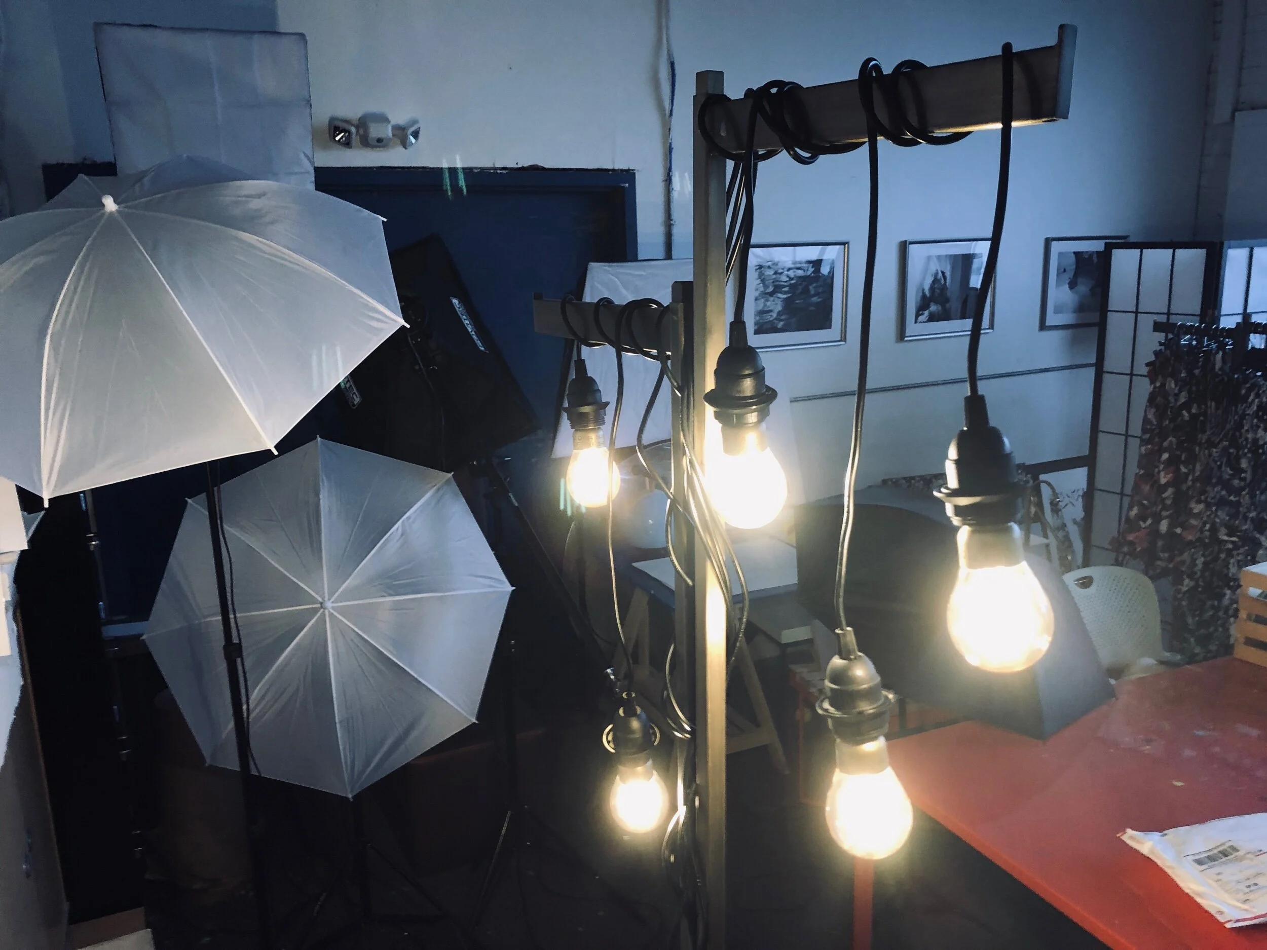 The image depicts a dimly lit room with several white umbrellas, multiple hanging black cables, and exposed light bulbs providing illumination. There are framed photos on the wall and a rack with clothes, with a table in the foreground holding papers