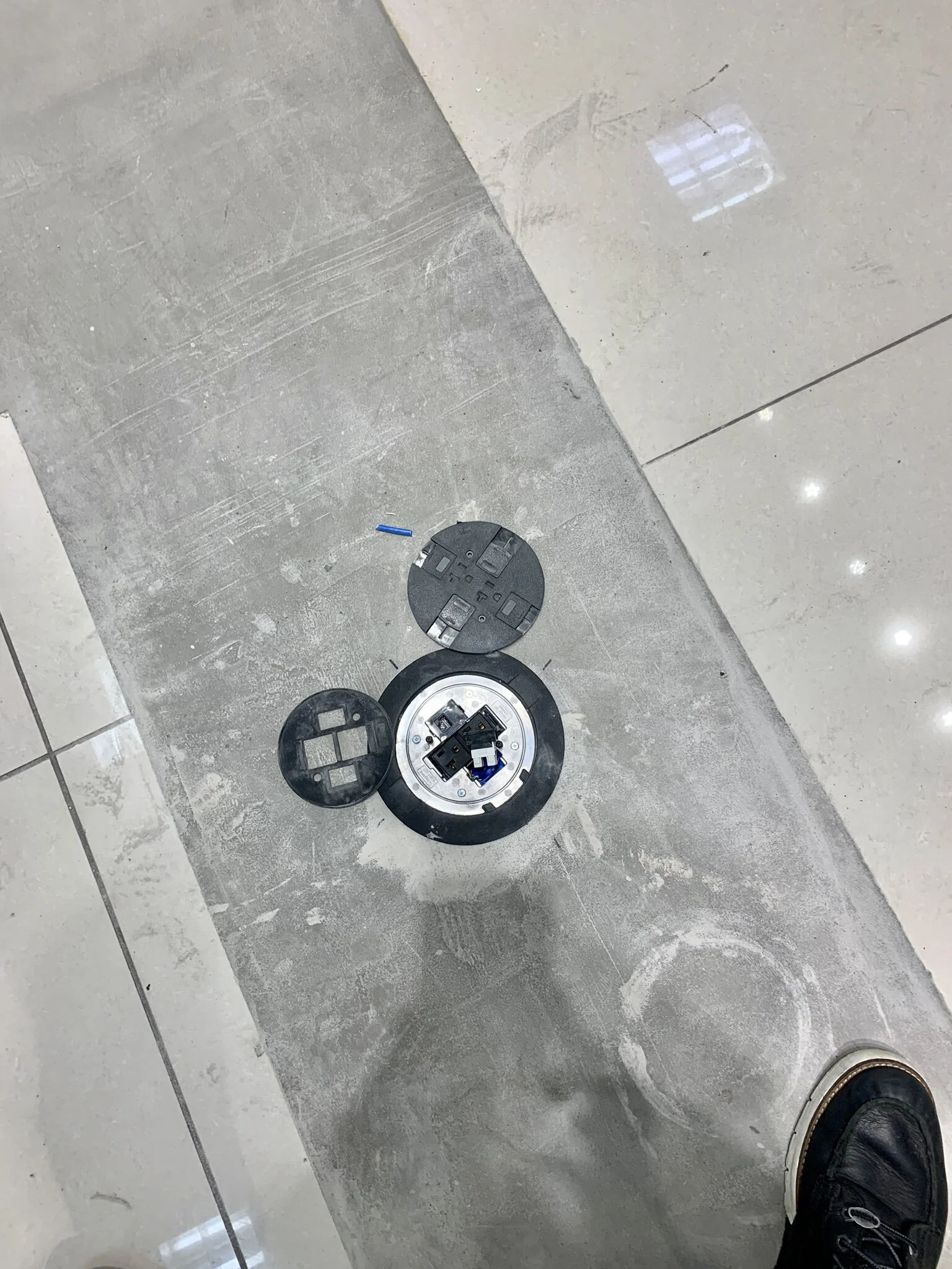 Disassembled electrical outlet or data port on a tiled floor, with a person wearing black shoes standing nearby.