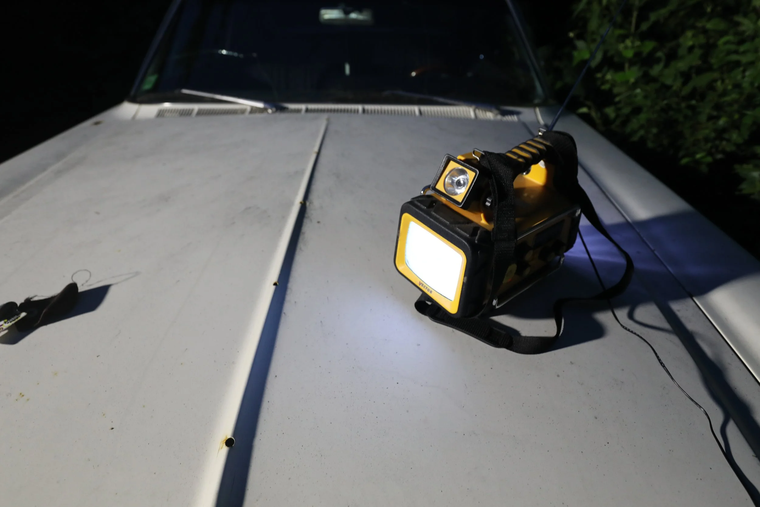 A flashlight positioned on the hood of a white vehicle during nighttime, illuminating the surface with bushes visible in the background.