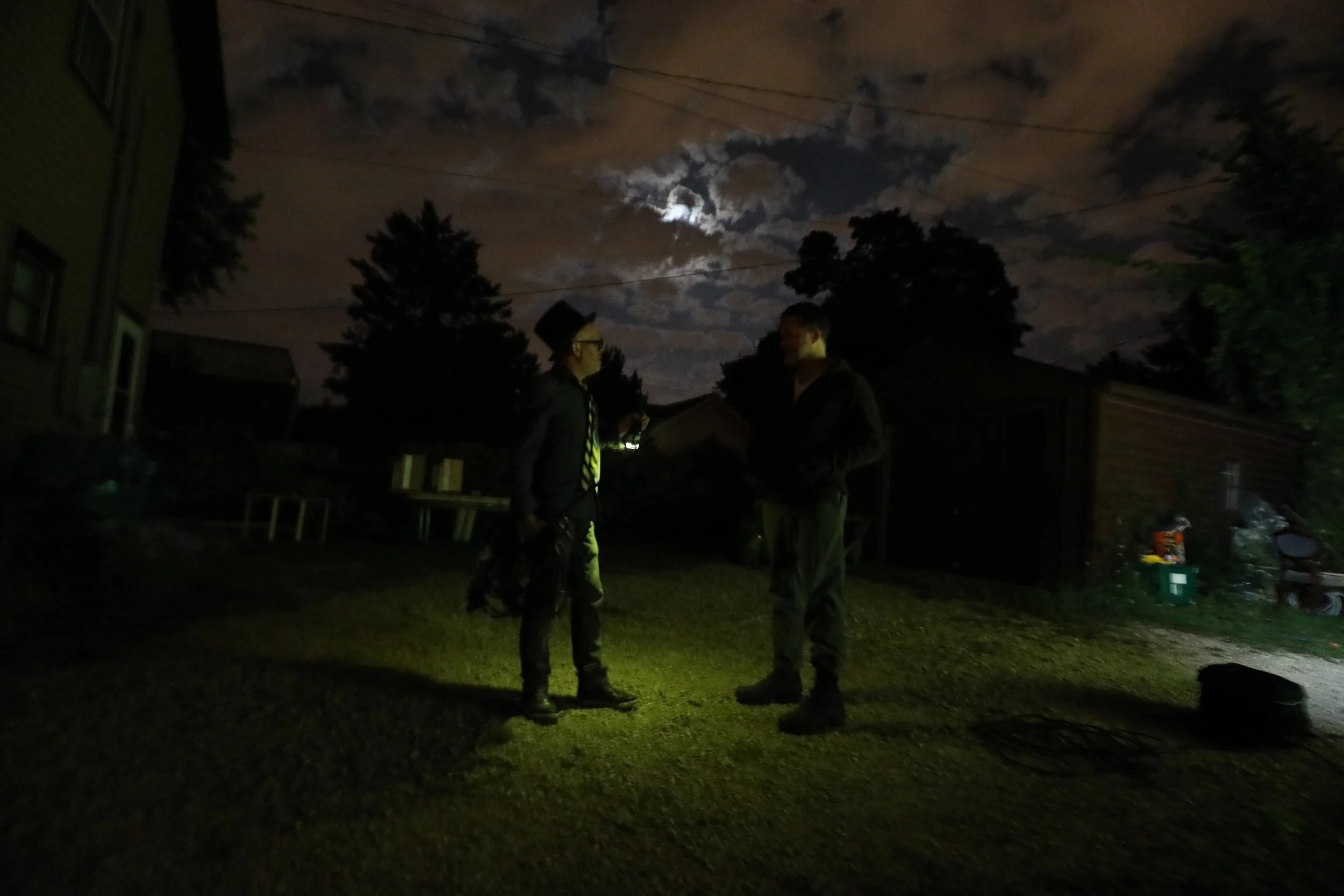 Nighttime backyard scene with two men talking, one holding a drink, trees, a house, and clouds with the moon in the cloudy sky.