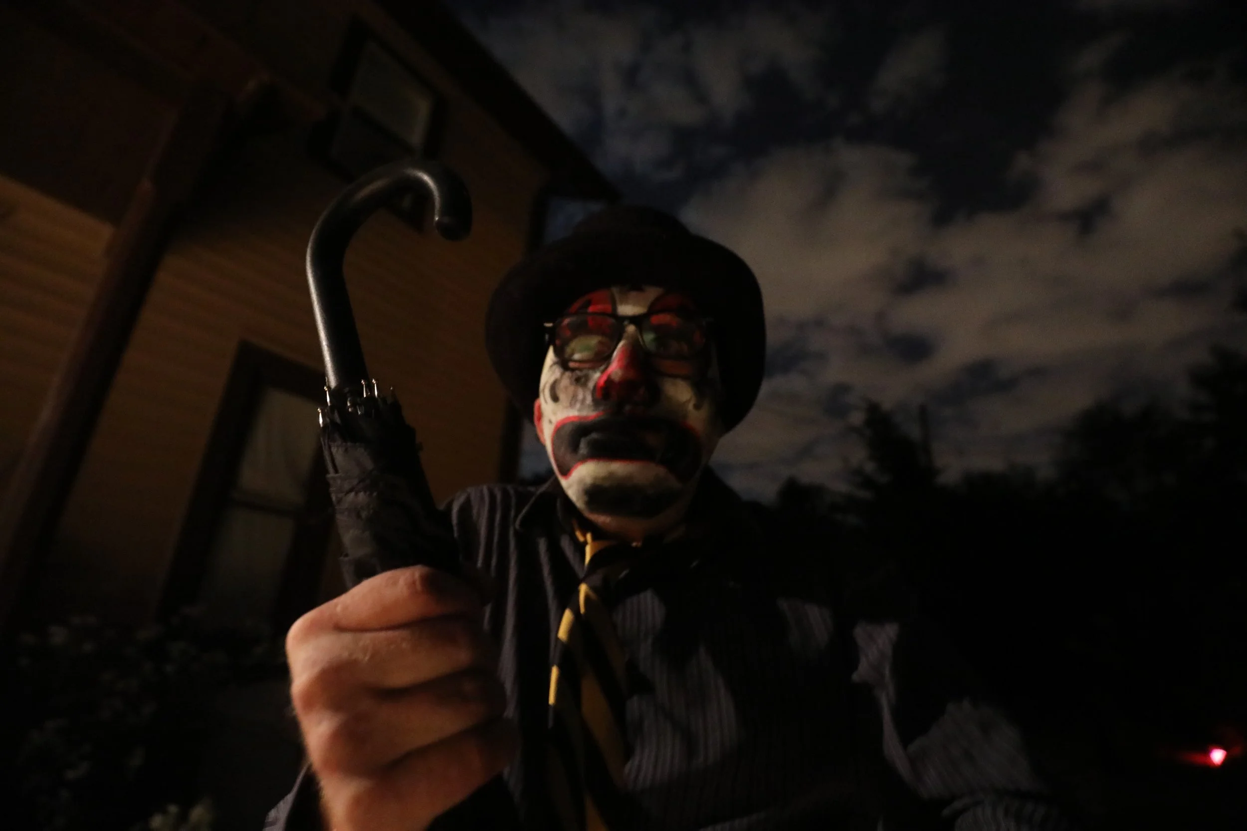 A person wearing face paint resembling a skeleton, glasses, and a black hat is holding an umbrella and taking a selfie outdoors at night with a dark sky and clouds in the background.