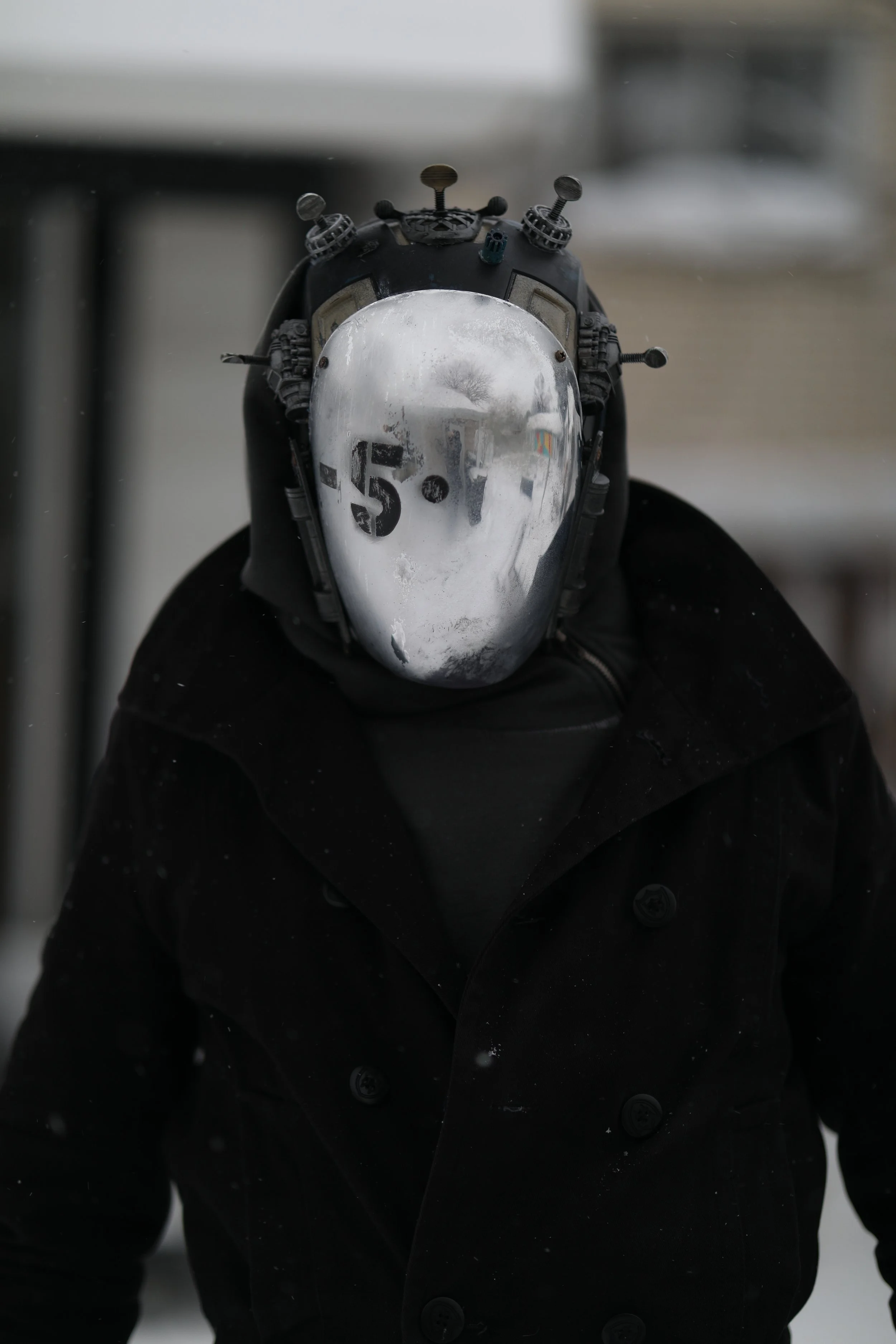 A person wearing a black coat with a reflective face mask, which has the number 5.1 and a percentage sign written on it, taking a mirror selfie outdoors in winter.
