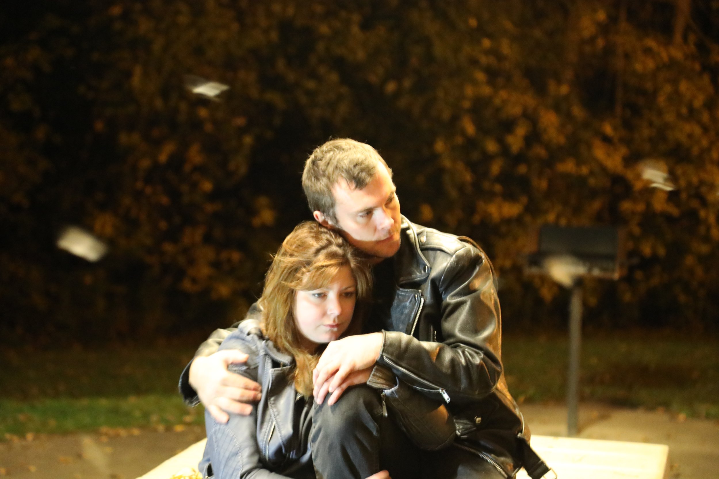 A man and a woman are sitting closely together outdoors at night. The man, wearing a leather jacket, is hugging the woman, who has brown hair and is wearing a denim jacket. The background is dark with blurred trees and some scattered leaves on the gr
