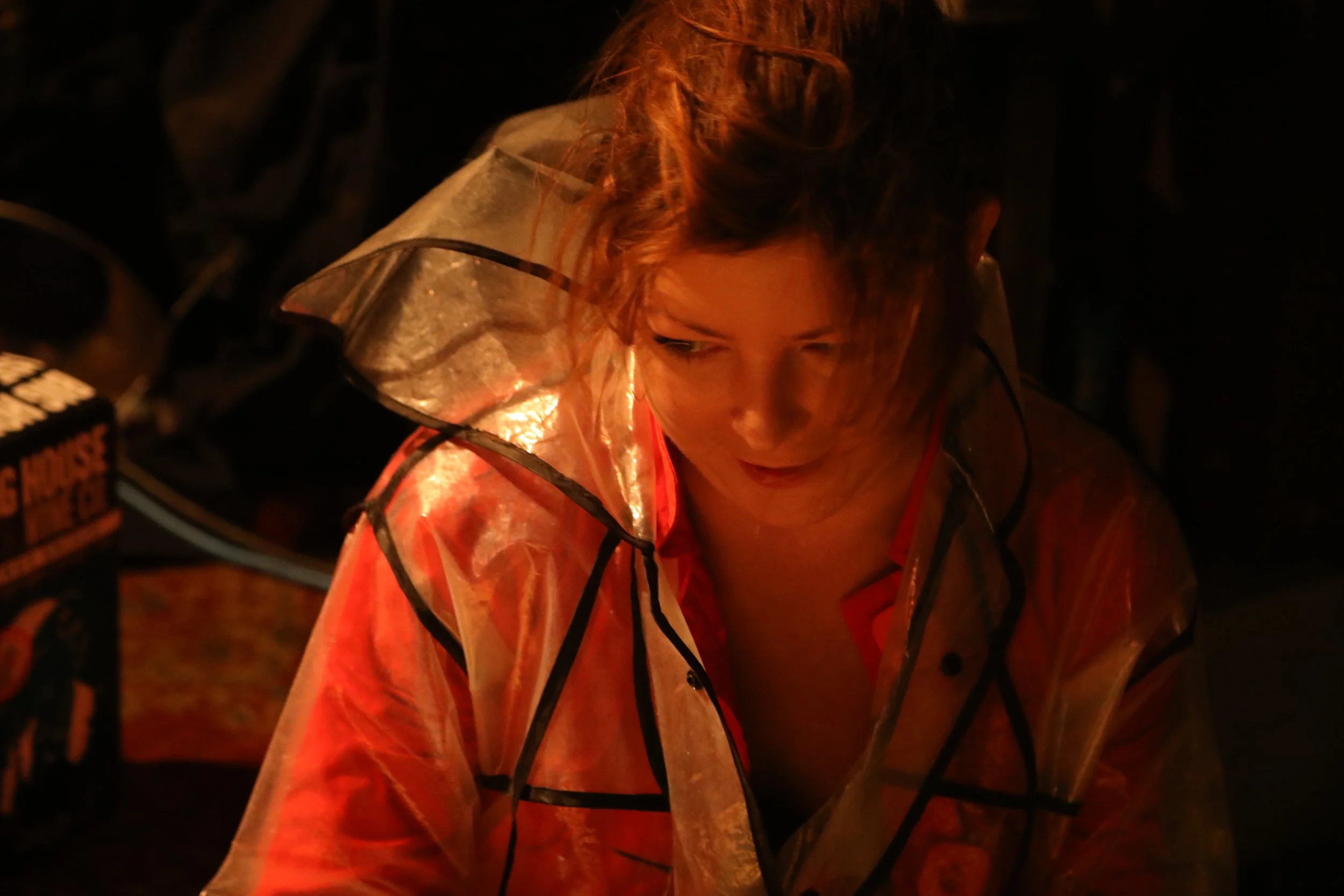 A person with curly hair is wearing a transparent raincoat with black trim and an orange shirt underneath. The scene is dark and lit by a warm, orange light.