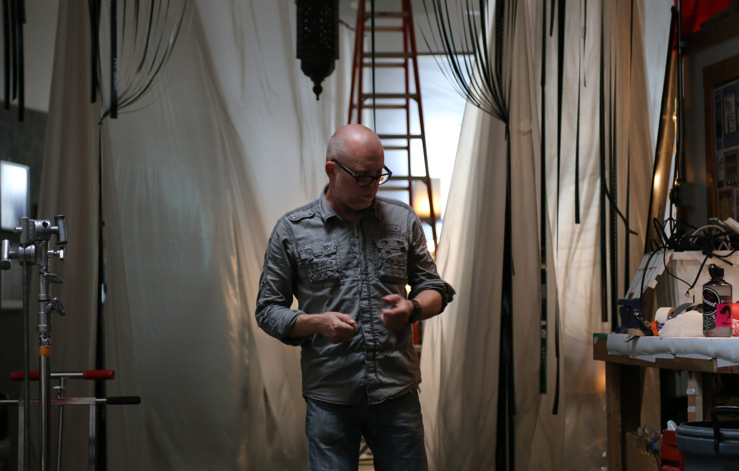 A man with glasses and a bald head standing in a workshop surrounded by equipment and tools.