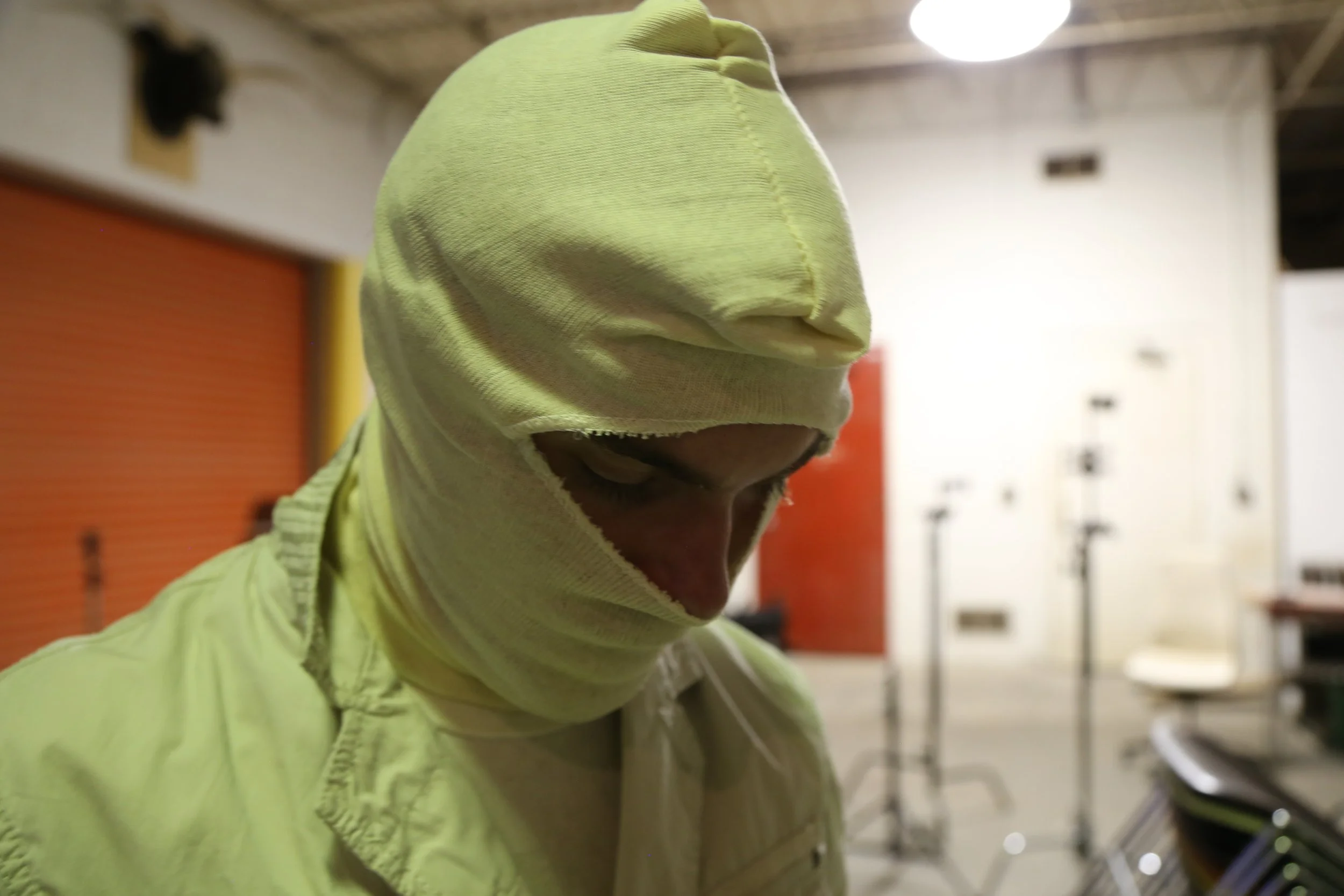 A person wearing a yellow hoodie with the hood pulled over their head, partially covering their face, in an indoor setting with tables and chairs in the background.