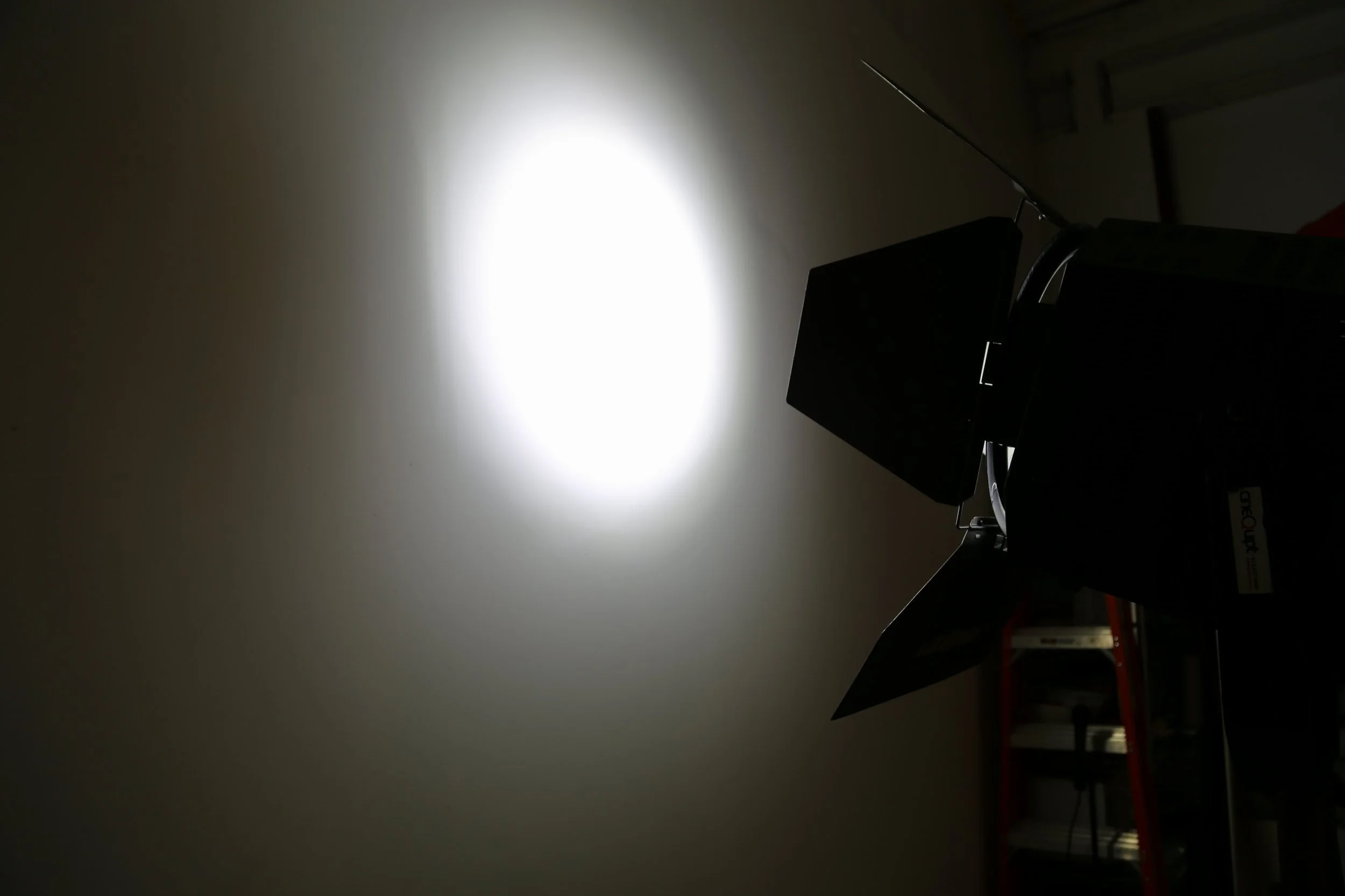 A projector mounted on a stand projecting light onto a white wall in a dark room with shelves in the background.