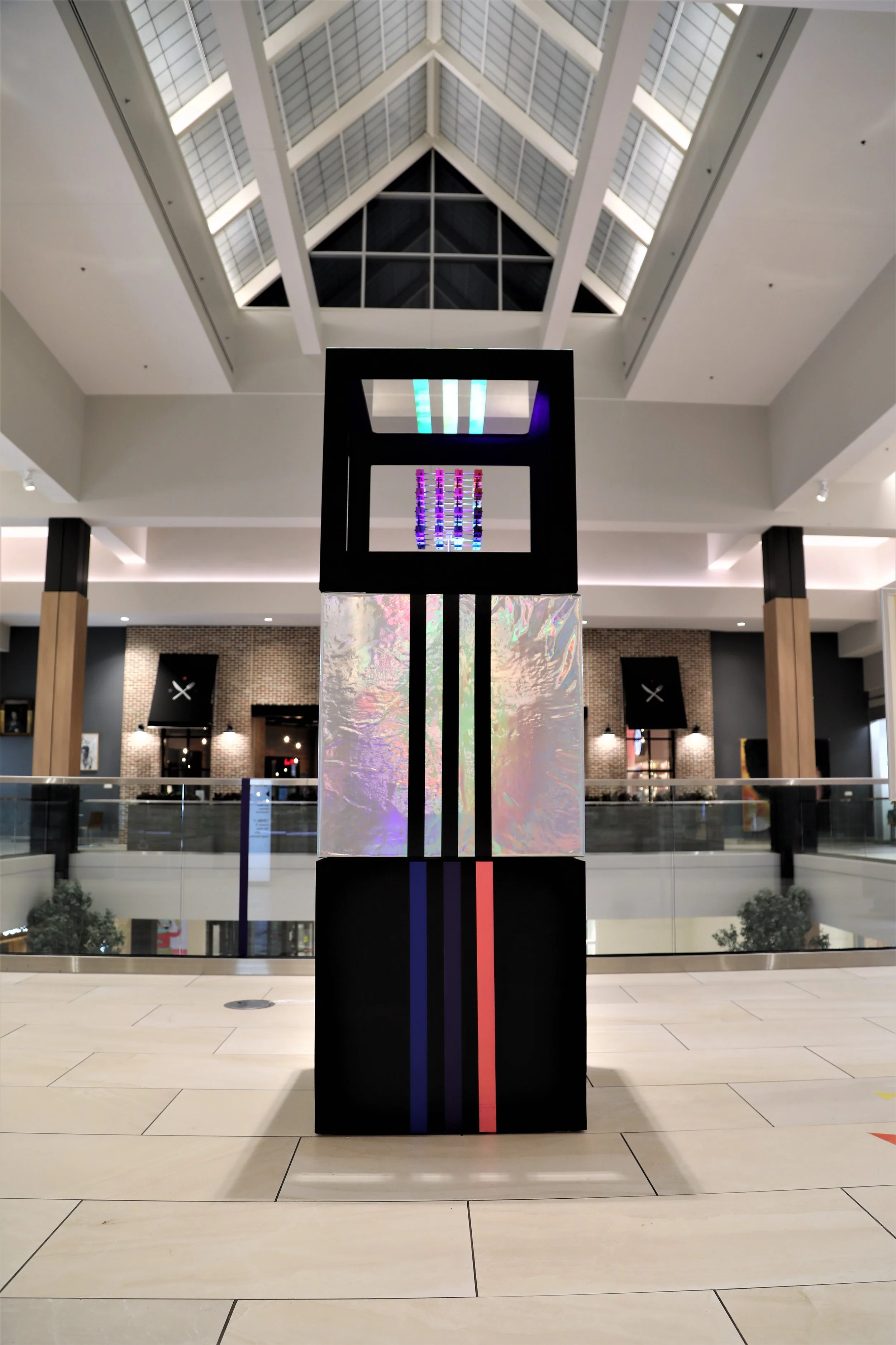 Modern art installation in a shopping mall with a tall black rectangular structure featuring vertical glowing lines of blue, green, and red lights. The background shows a spacious, well-lit atrium with a glass ceiling and second level shops.