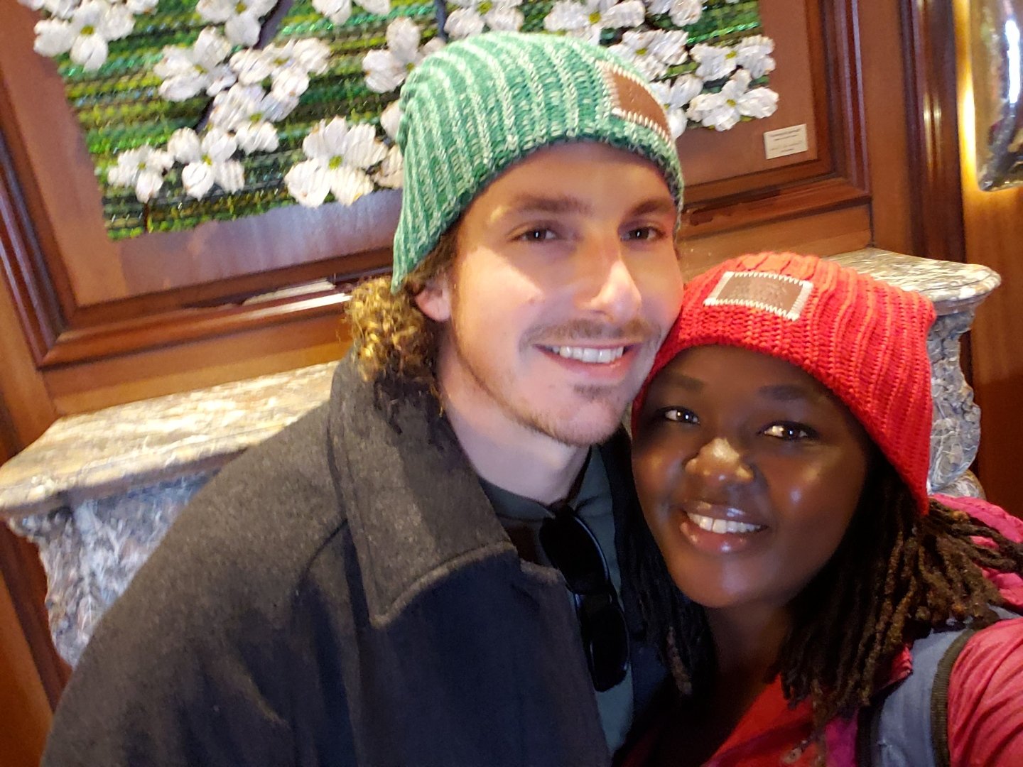 A couple wearing winter hats poses for a selfie indoors, in front of a decorative floral artwork.