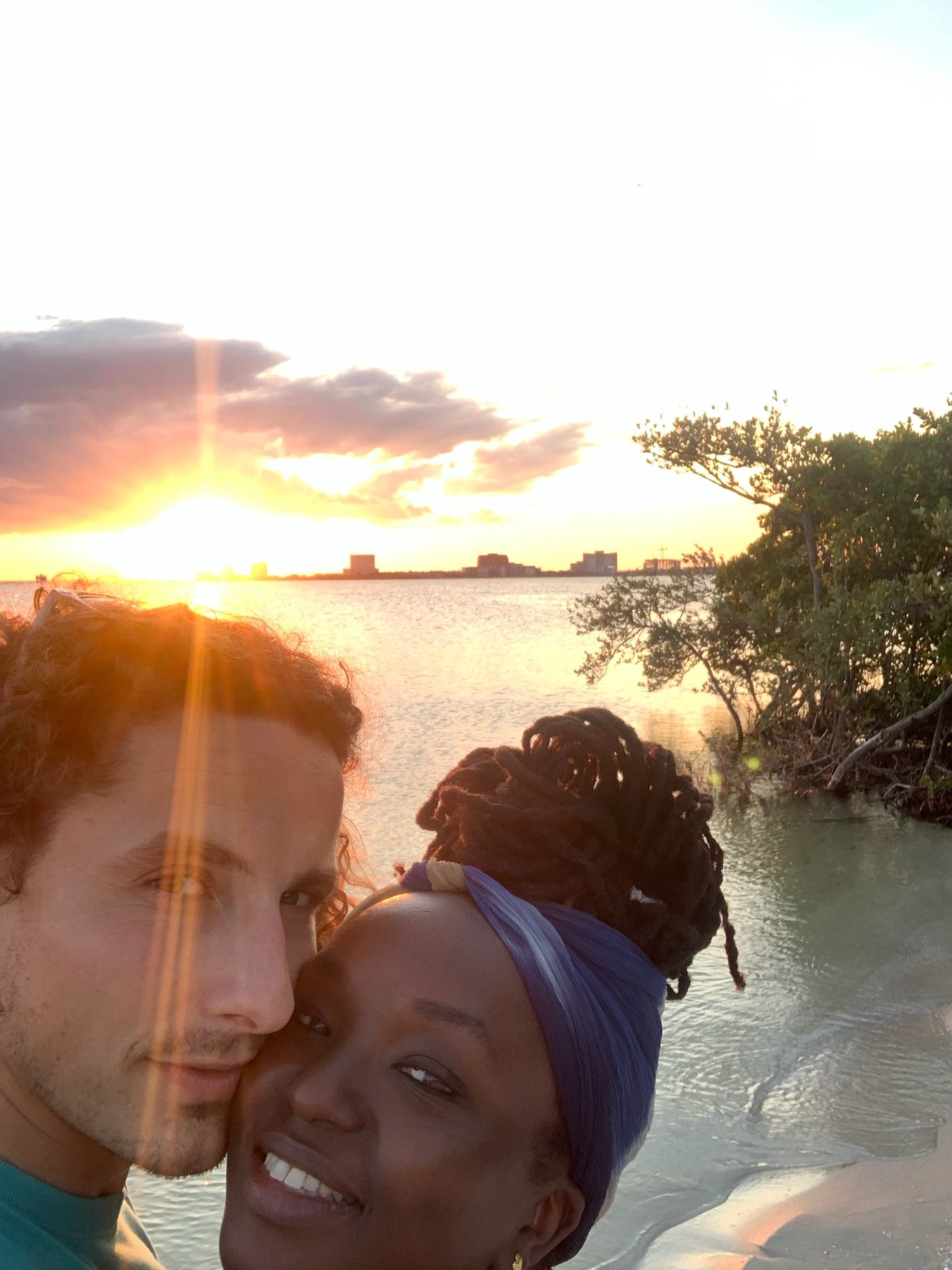 Couple taking a selfie at the beach during sunset, with water and a distant skyline in the background.