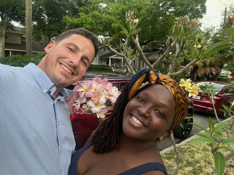 Two people taking a selfie in front of flowering trees and cars on a residential street.