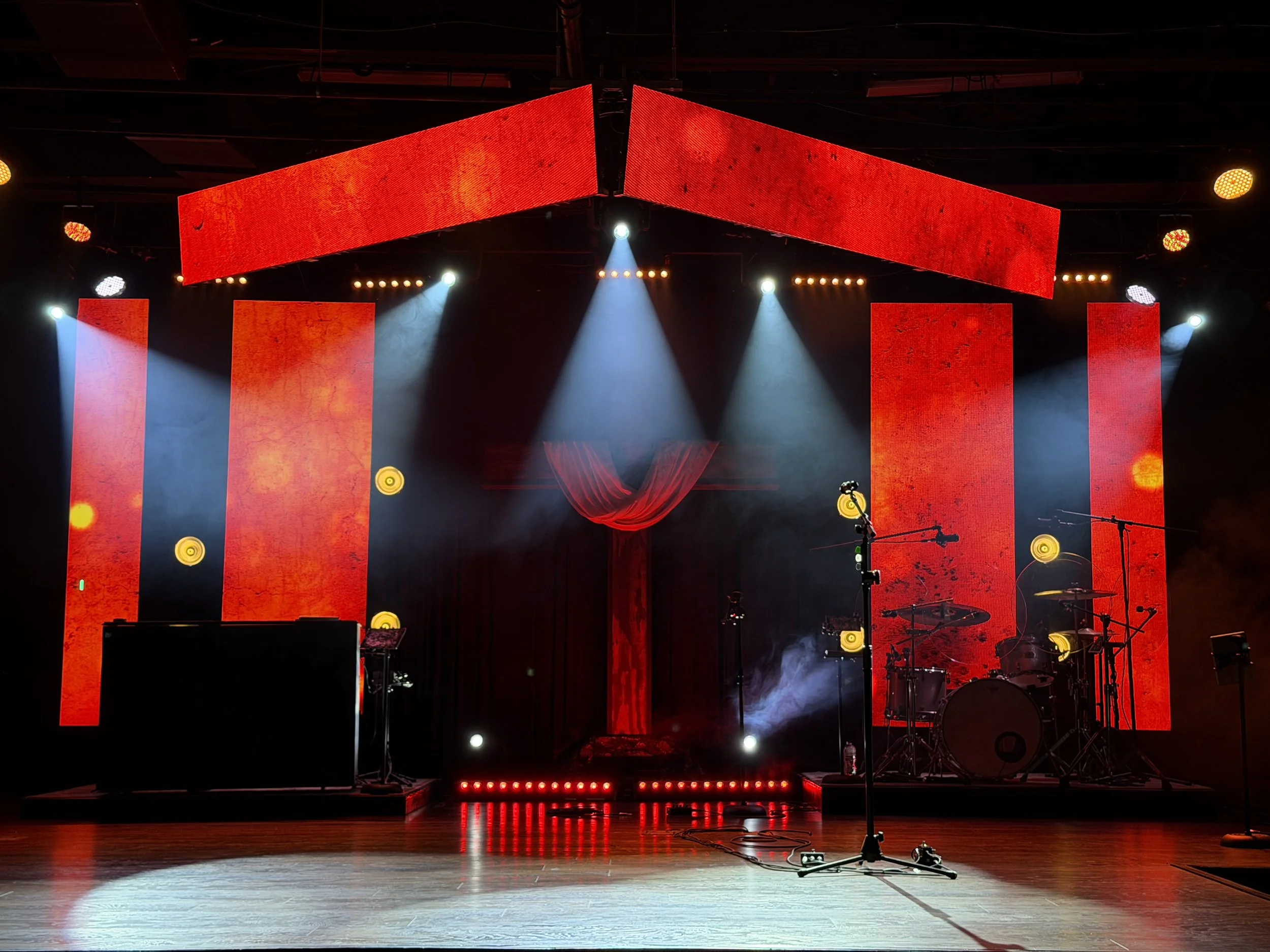 Empty stage with large red LED screens, spotlights, musical instruments including drums, microphone stands, and stage equipment, illuminated by red and white lights.