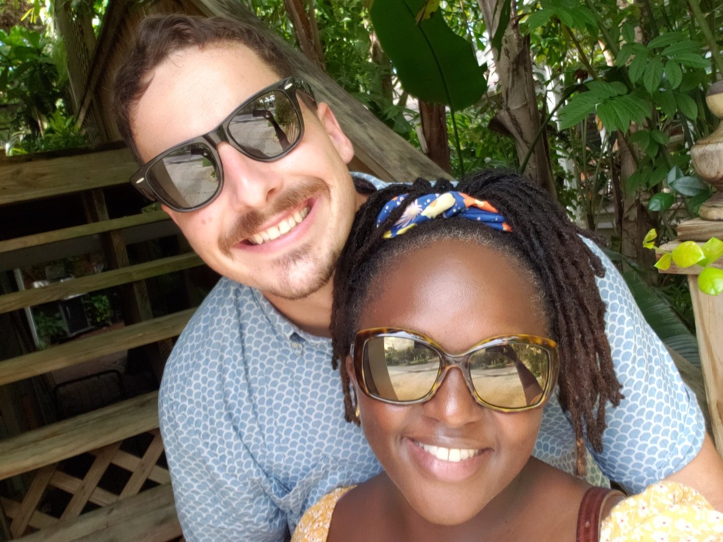 A man and woman smiling together wearing sunglasses, with green foliage and wooden railing in the background.