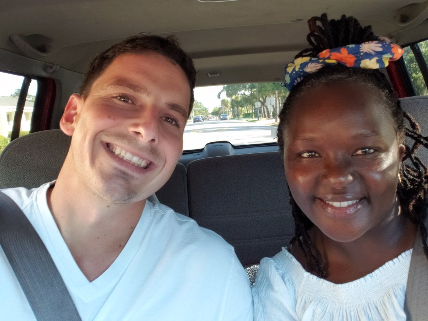 Two people smiling in a car selfie, wearing casual clothes, with seatbelts fastened.