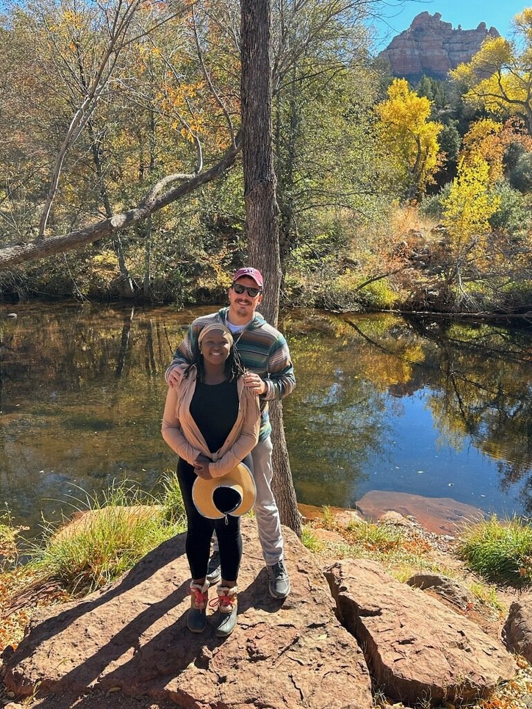 Two people standing on a rocky area near a river with autumn trees in the background.