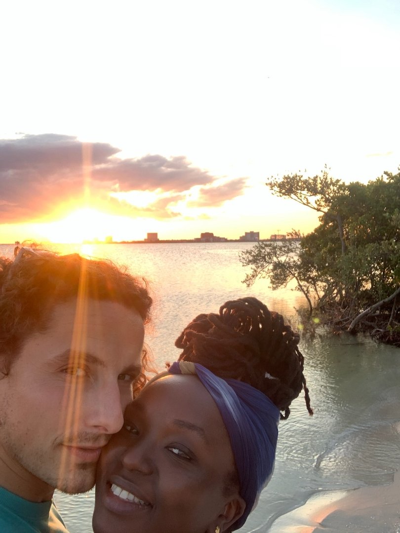 A smiling couple taking a close-up selfie on a beach during sunset. The sun is setting on the horizon, casting a warm glow on the water and clouds. Trees and shrubs are visible on the sandy shore.