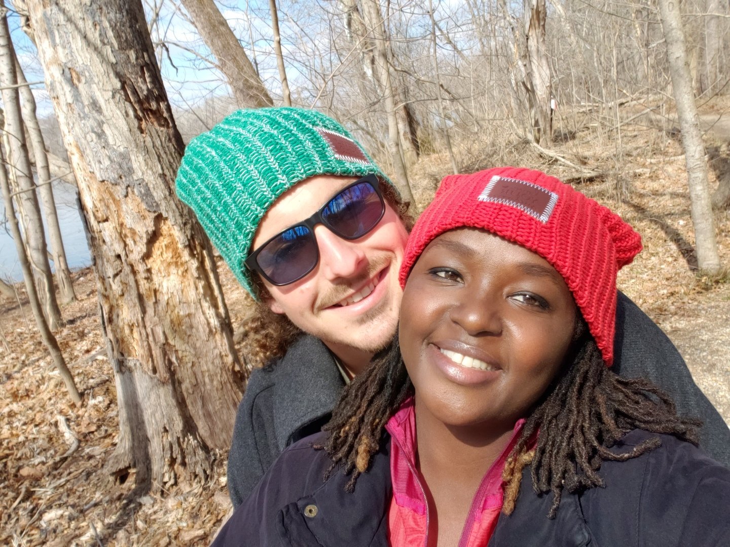 Two people smiling, wearing colorful knit beanies, standing in a wooded area during fall or winter.