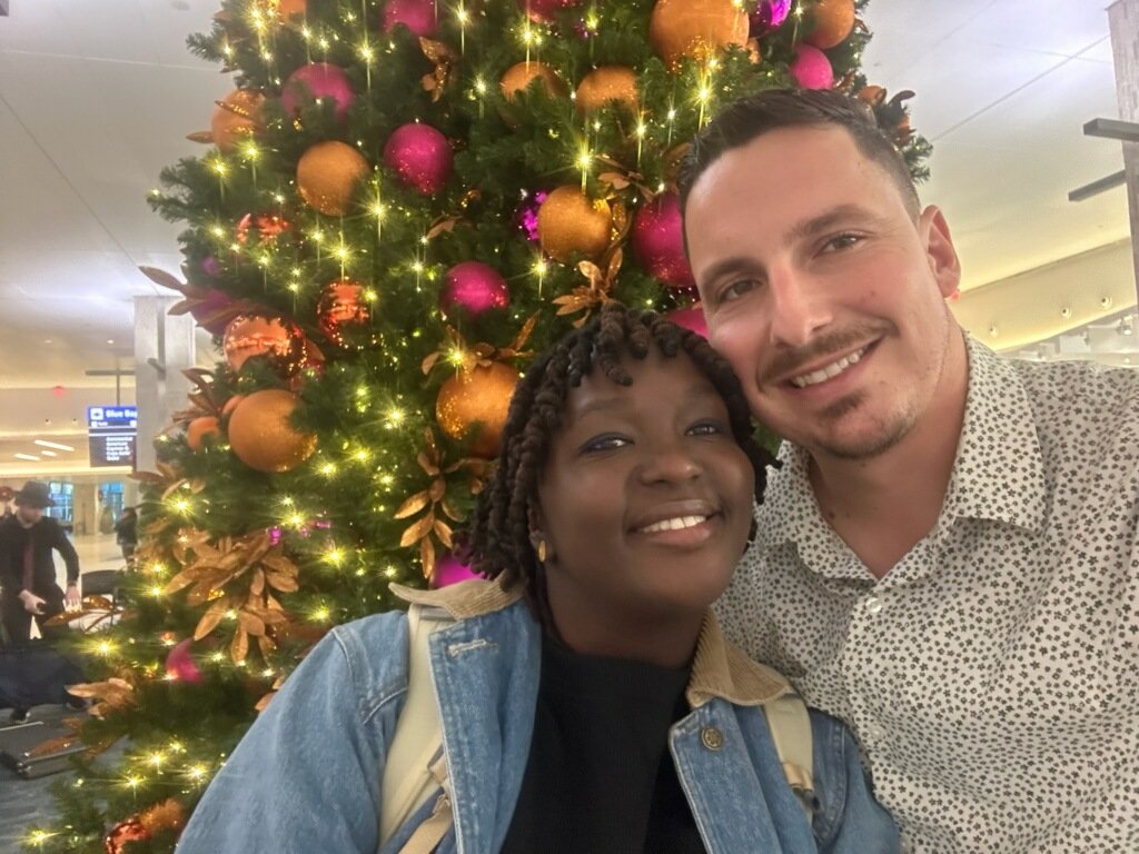 A smiling couple takes a selfie in front of a decorated Christmas tree adorned with orange and pink ornaments and lights inside an airport terminal.